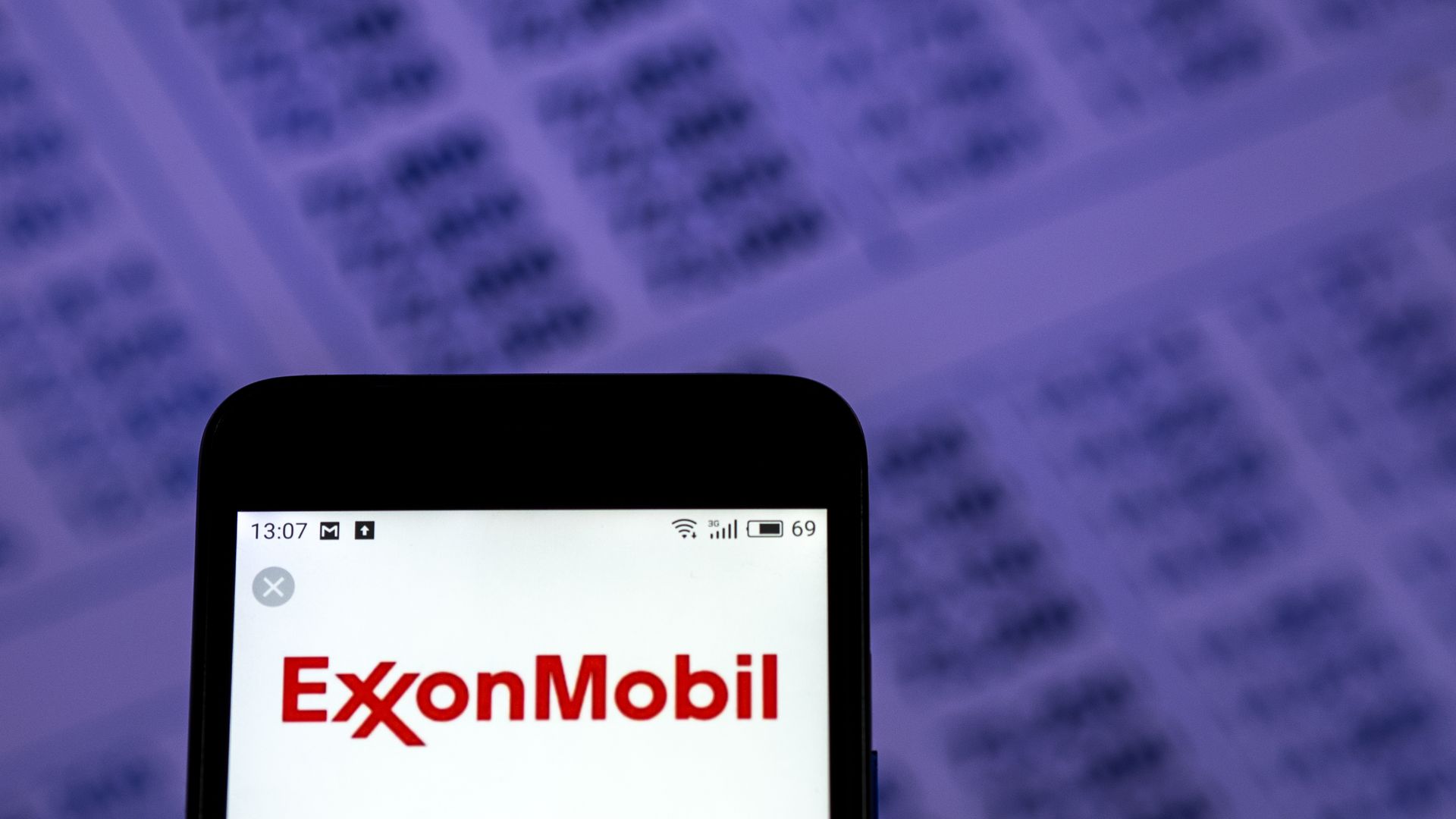 In this image, a phone displaying the ExxonMobil logo is displayed in front of a purple background with numbers.
