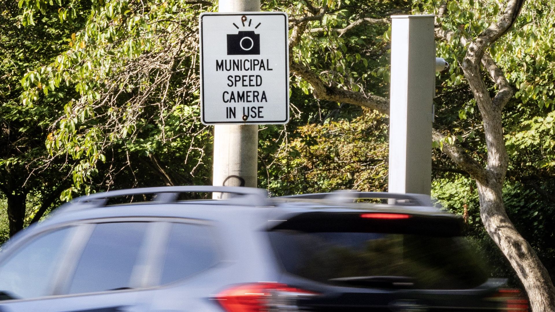 A car zooms by a sign that reads "Municipal Speed Camera In Use"