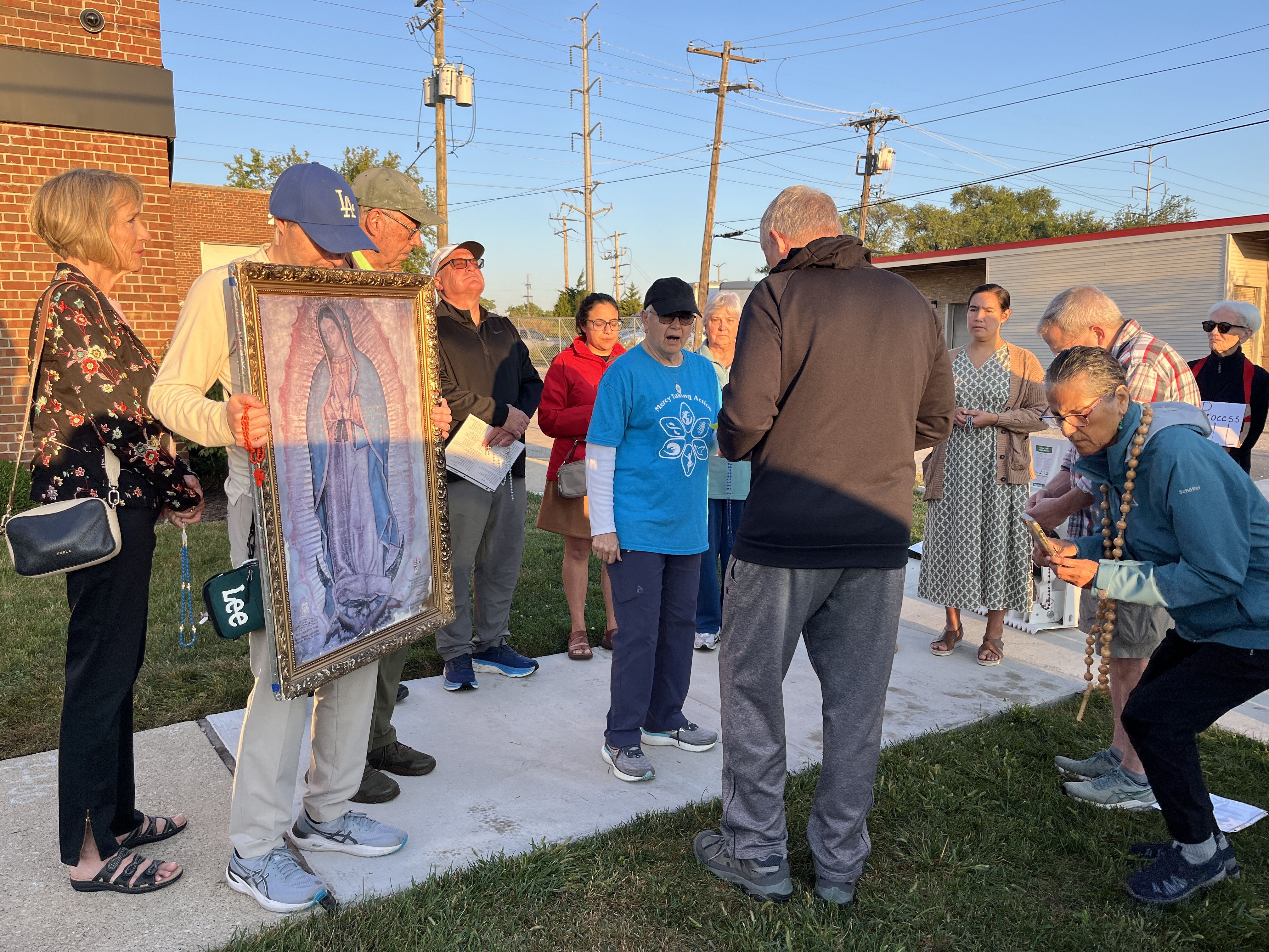 Group of people standing outside near a brick building and utility poles, some holding rosaries and a framed image of the Virgin of Guadalupe, engaged in a prayer or gathering at sunset.