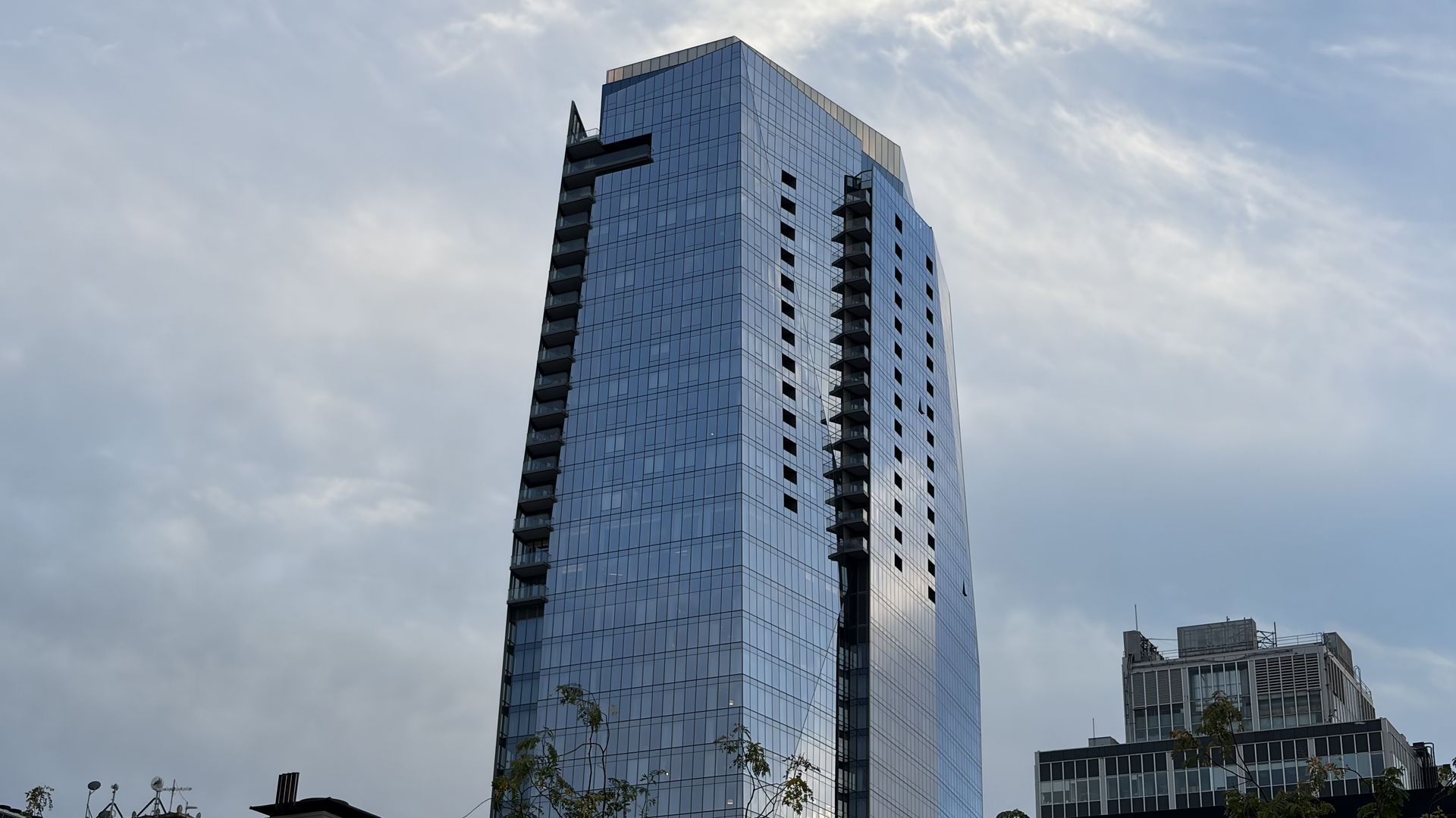 Tall glass skyscraper with blue reflective windows and narrow balconies along both sides, rising against a cloudy sky; lower buildings and trees visible at the bottom.