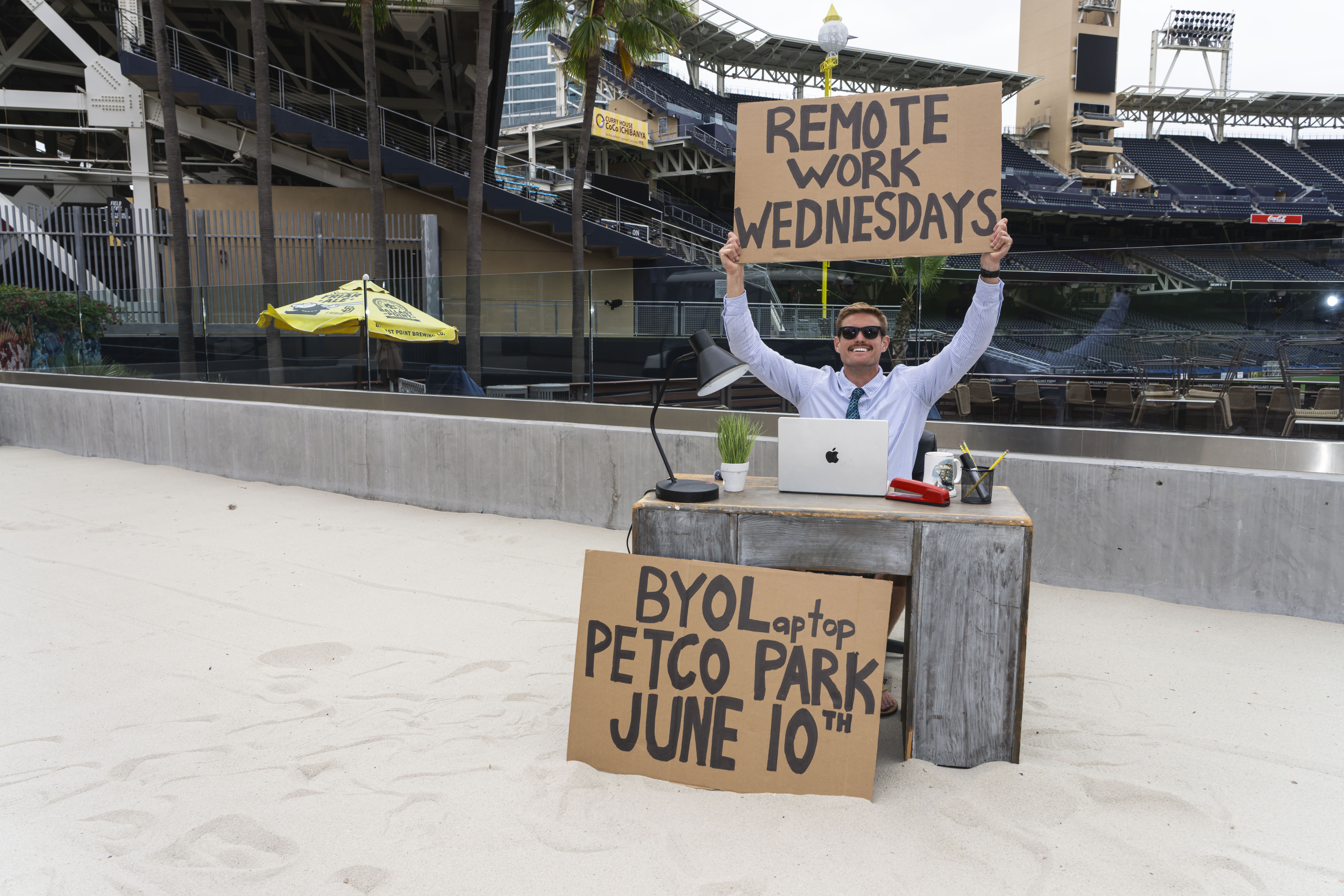 A man wearing sunglasses and a striped shirt sits at a desk on a sandy baseball field, holding a large cardboard sign that reads "REMOTE WORK WEDNESDAYS"; laptop, lamp, and plant on the desk; empty stadium seats in the background.