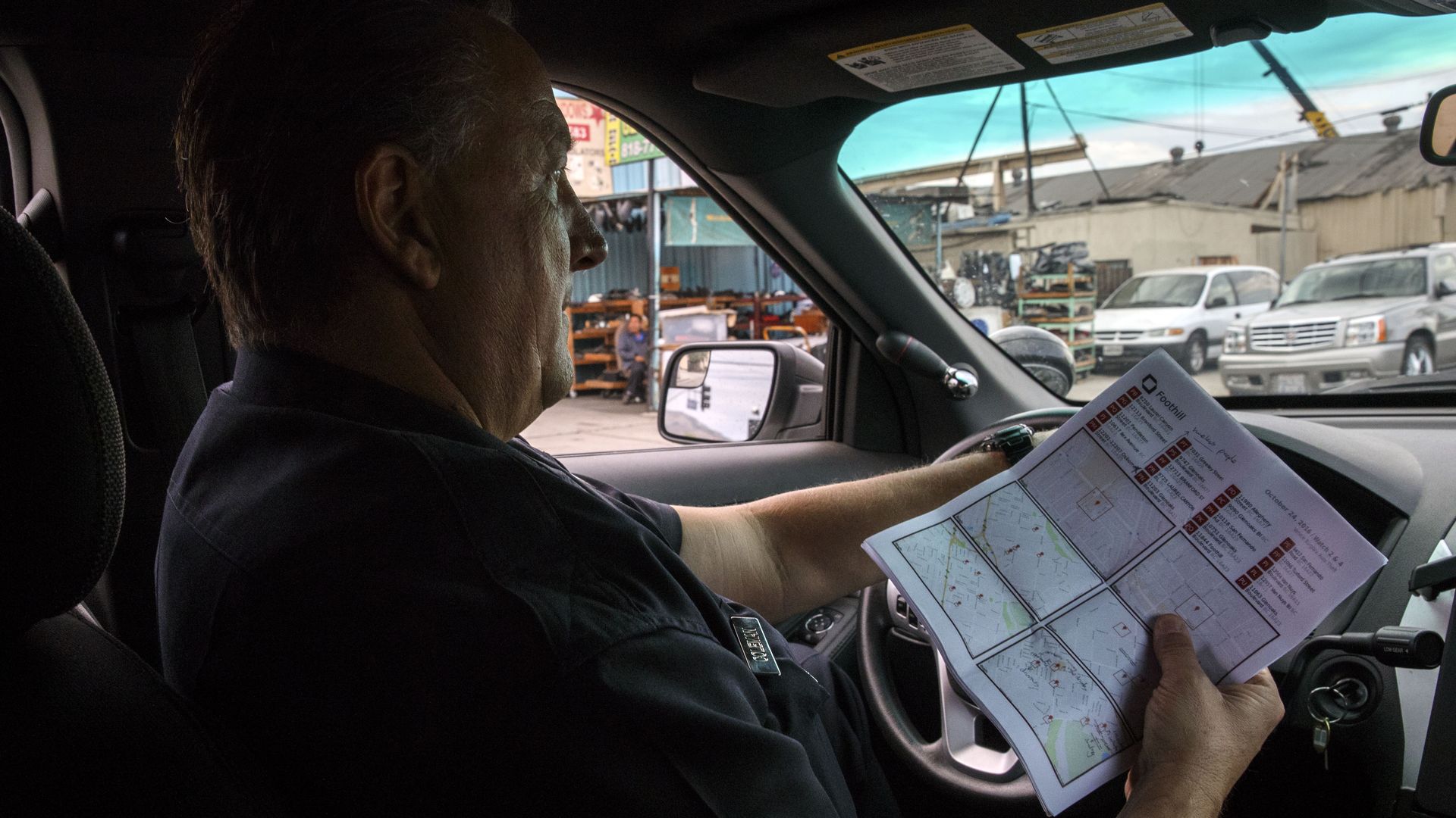 A police officer drives a car while holding a piece of paper with several maps on it