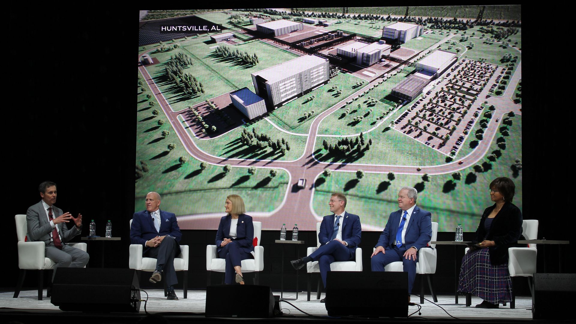 Five panelists in business attire seated on stage discussing with a large screen behind showing an aerial view of a building complex labeled "Huntsville, AL."