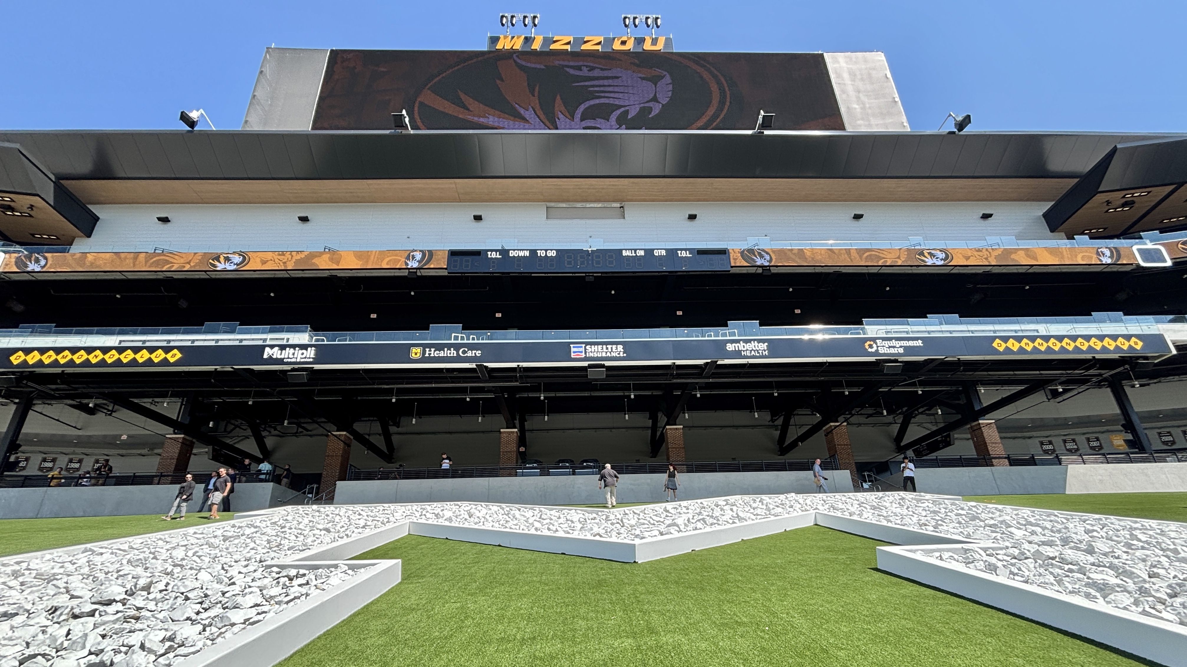 Exterior view of Mizzou stadium with orange-and-black branding, a large tiger logo on a dark backdrop, a scoreboard, and a few people walking the concourse under a blue sky.