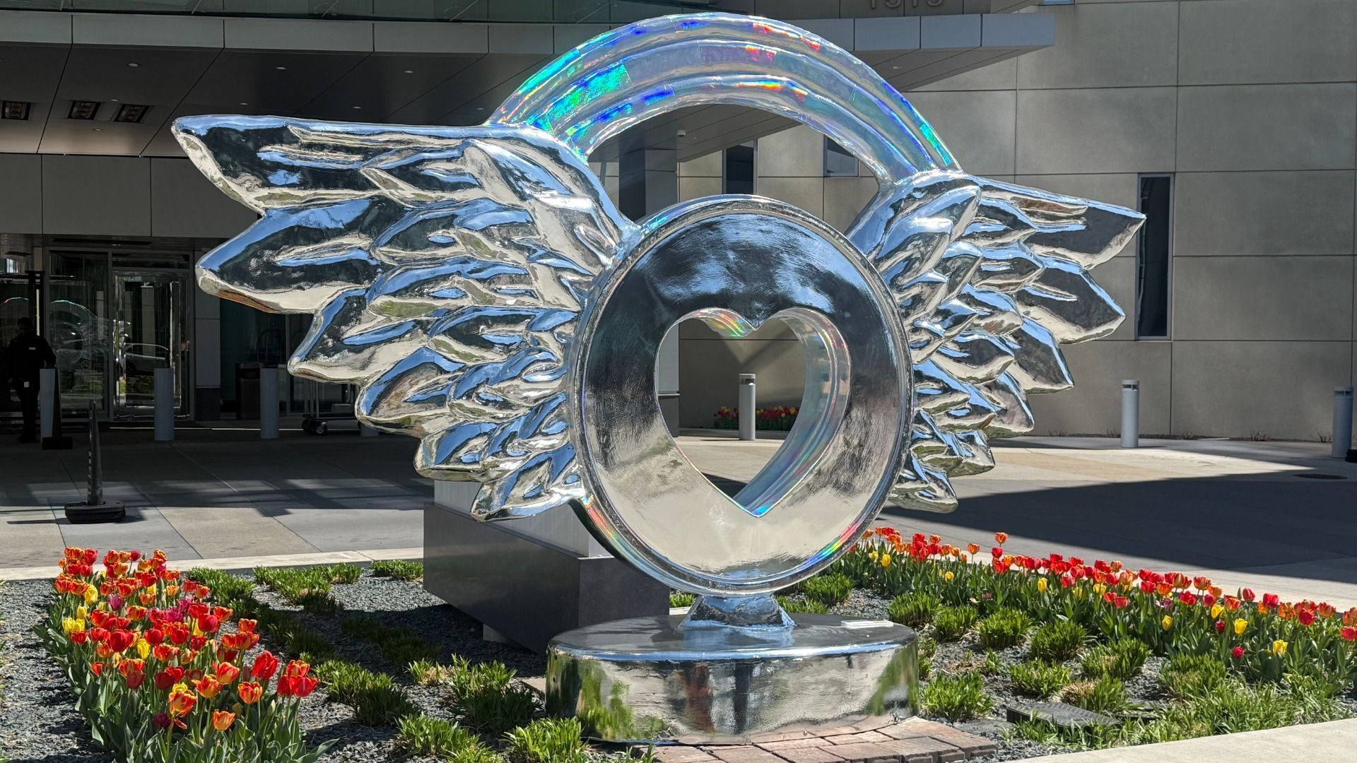 Shiny metal sculpture with wings on each side and a circular ring with a heart-shaped opening, topped by a rainbow arc. It stands before a modern building, surrounded by red and yellow tulips.
