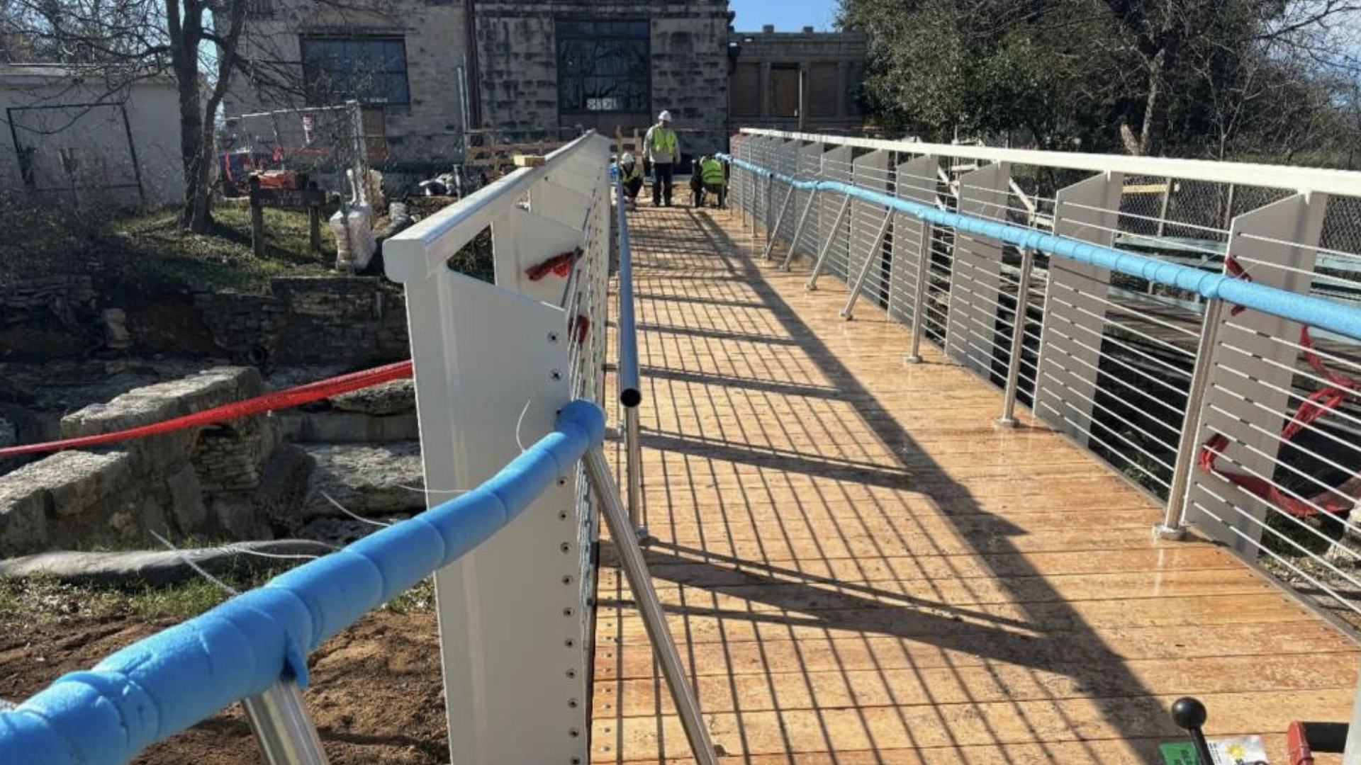 A newly constructed wooden pedestrian bridge with steel railings covered in blue protective foam, casting shadows, with three workers in green safety vests at the far end under a clear sky.