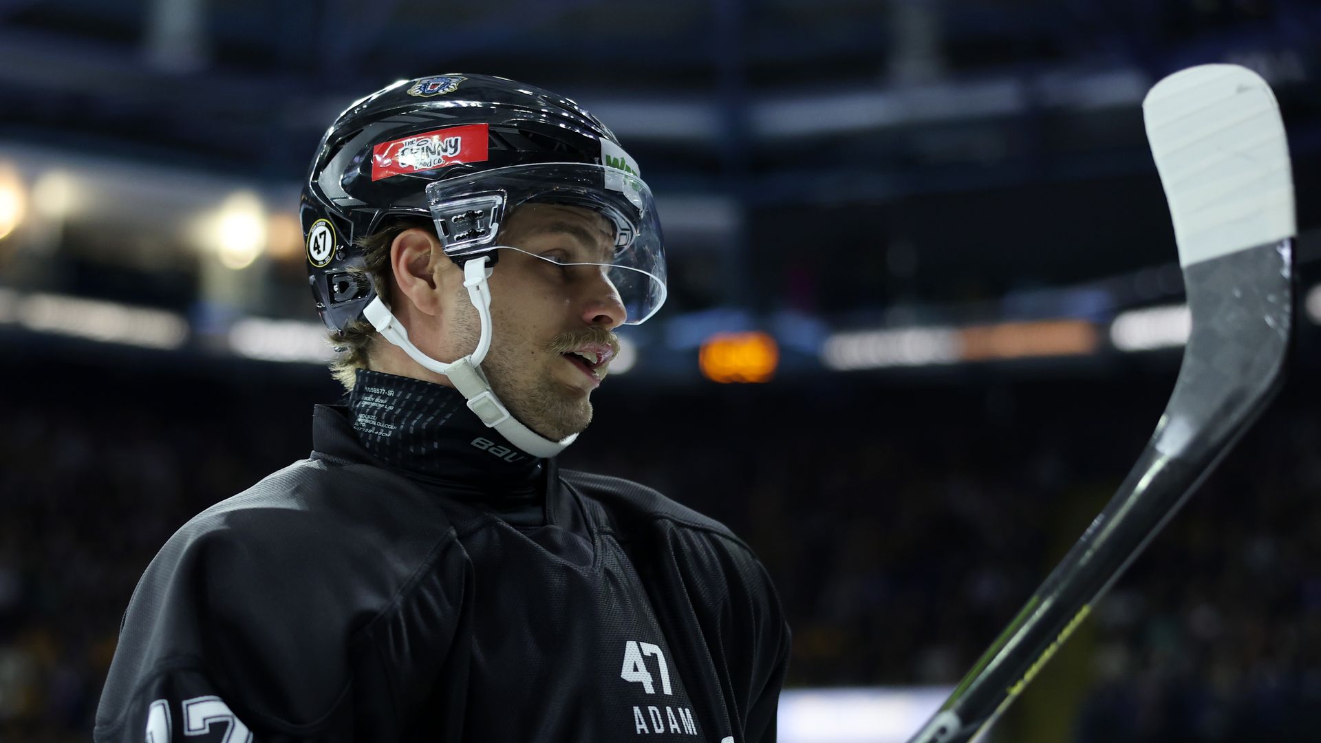 A hockey player with his taped stick raised in the air wears a black jersey, a helmet, and a black neck guard