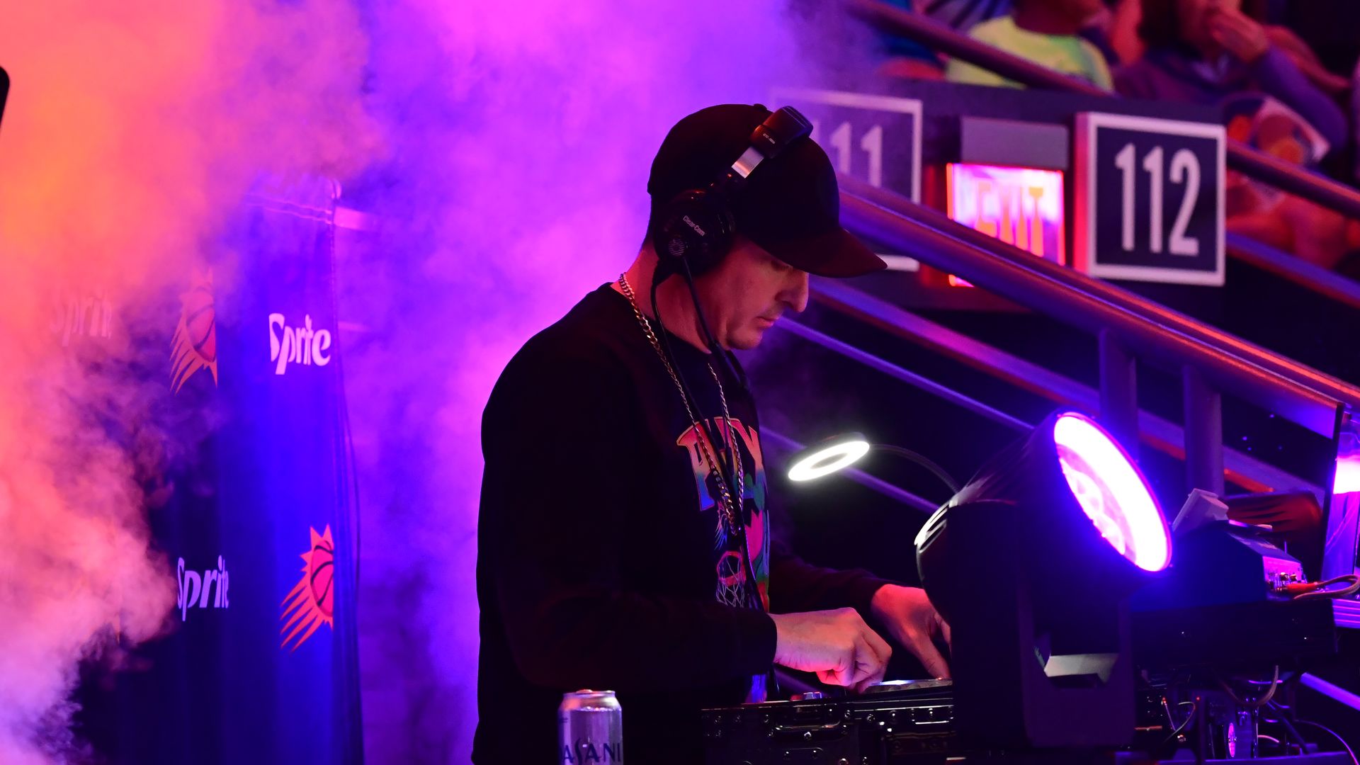 A man wearing headphones pushes buttons on a panel while surrounded by smoke illuminated by purple and orange lights.