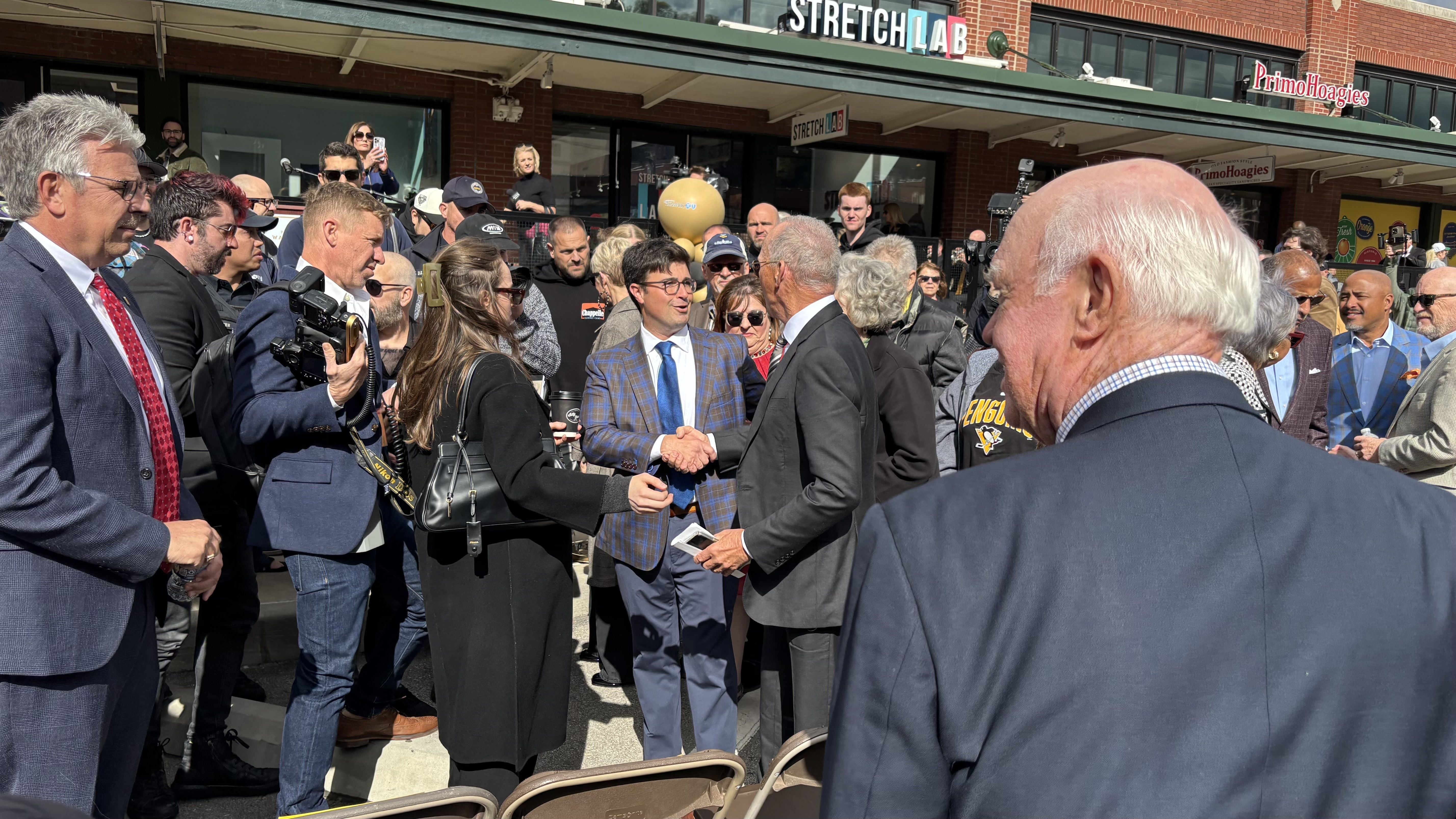 A group of people, some in suits, gathered outside a building with signs for "StretchLab" and "PrimoHoagies." Two men in the center are shaking hands, surrounded by photographers and onlookers.
