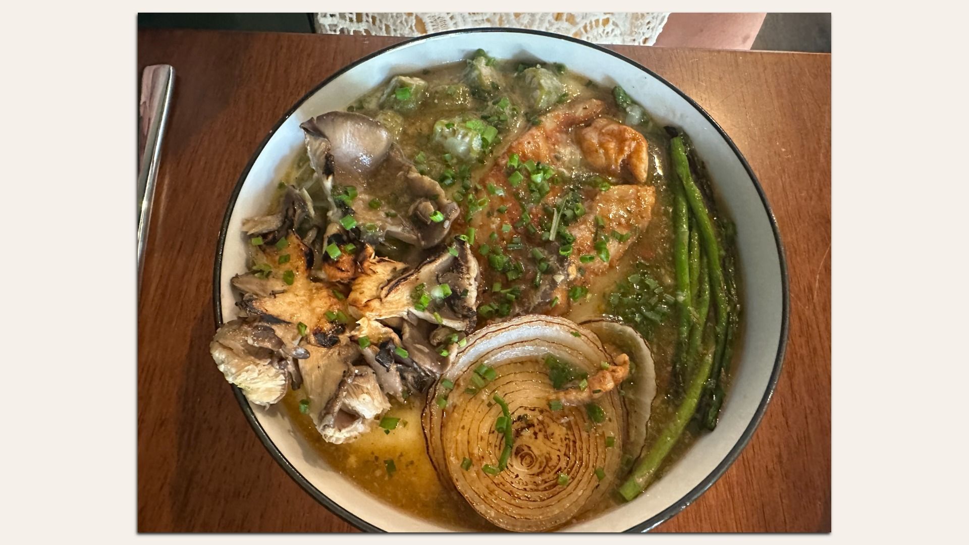 Top-down view of a white bowl with a dark rim, filled with a brown broth, pork slices, chopped green onions, a large onion ring, and long green vegetables on a wooden table with a nearby utensil.