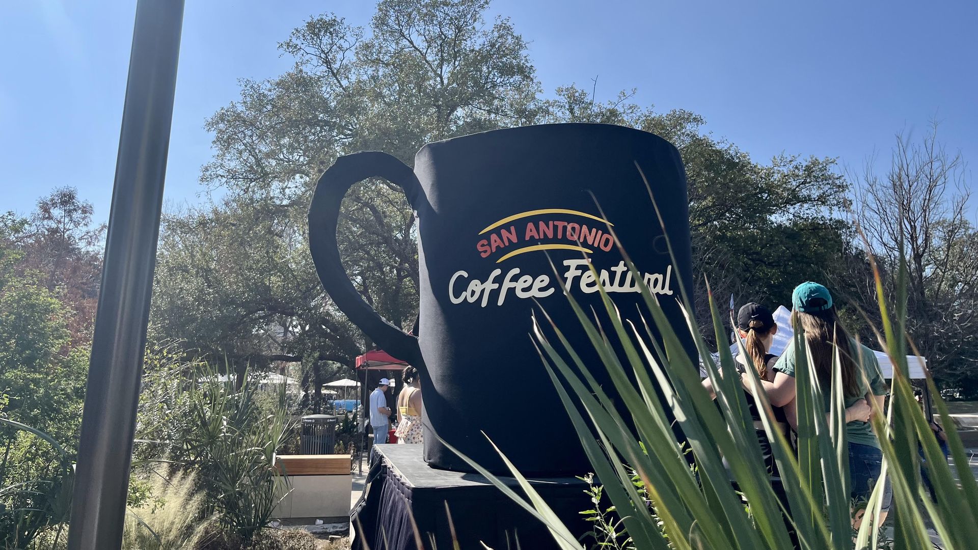 A large black coffee cup sculpture with "San Antonio Coffee Festival" on it sits in the middle of Civic Park in February 2025. It shows a sunny day with trees and people in the background.