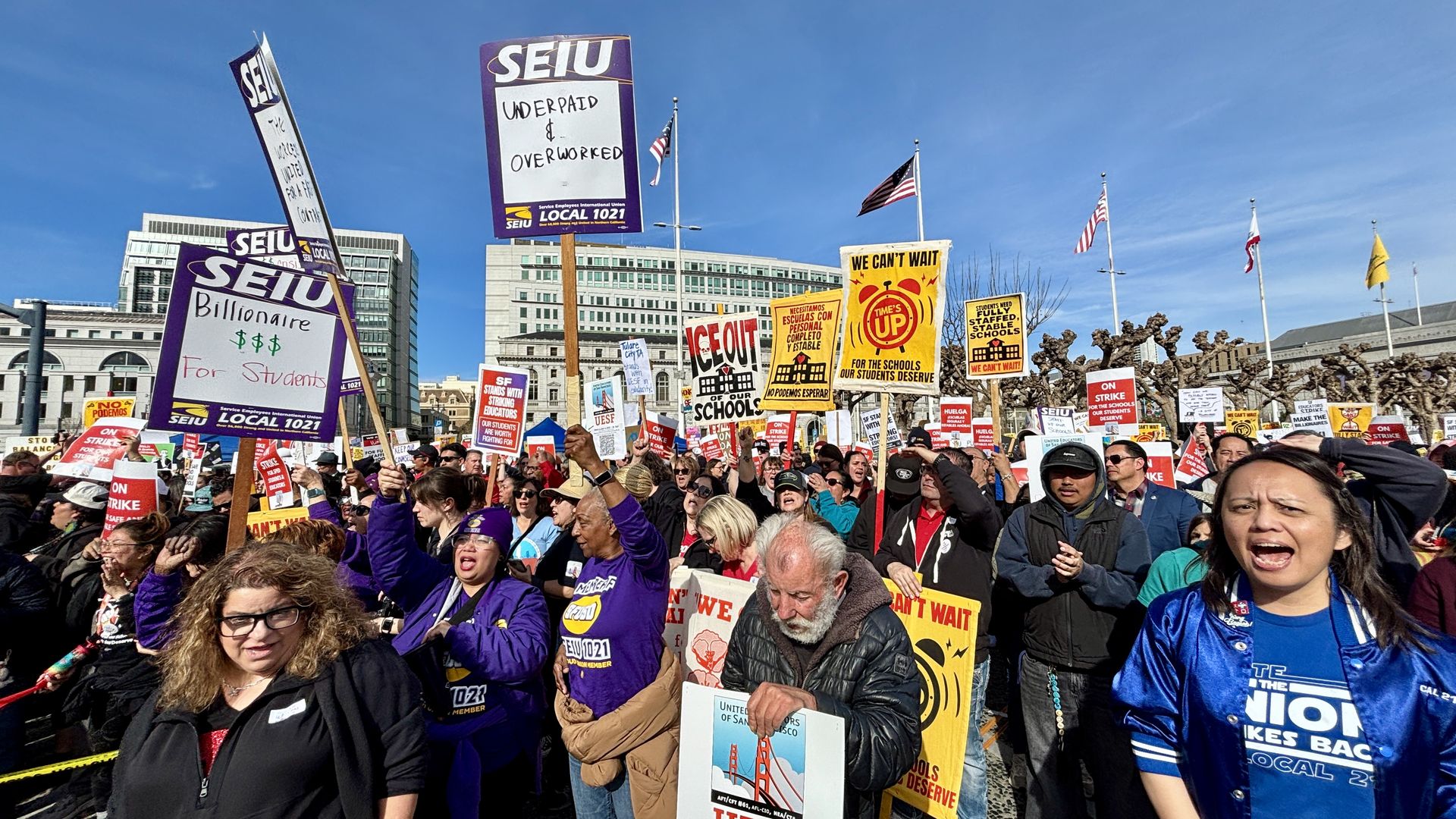 People at a protest in front of a large classical building, with yellow and red banners reading "WE CAN'T WAIT" and signs saying "ON STRIKE FOR OUR STUDENTS".
