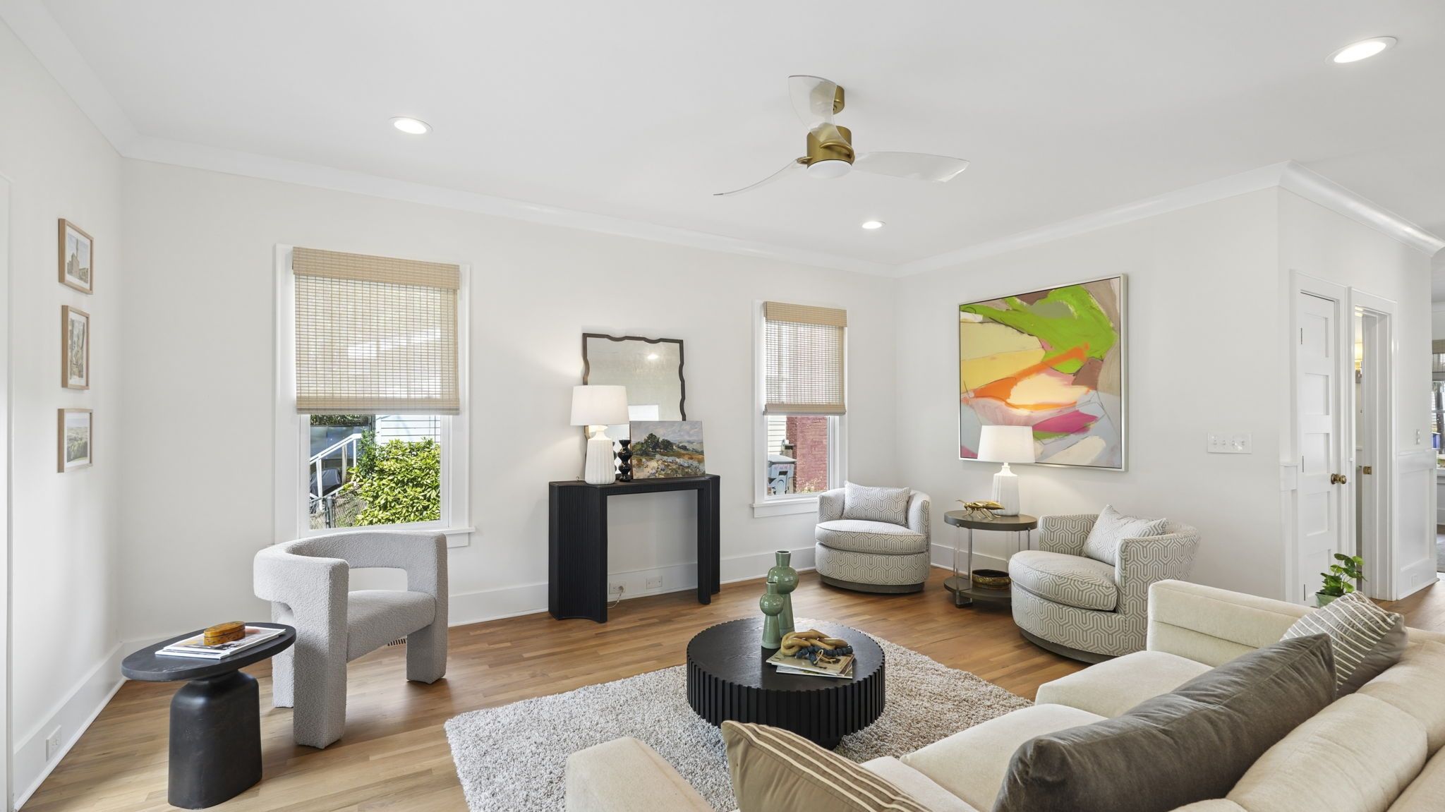 Bright living room with white walls, light wood floors, and a ceiling fan. Two windows with blinds; a black console with a mirror and lamps; circular chairs; beige sofa; black coffee table on a cream rug; abstract art.