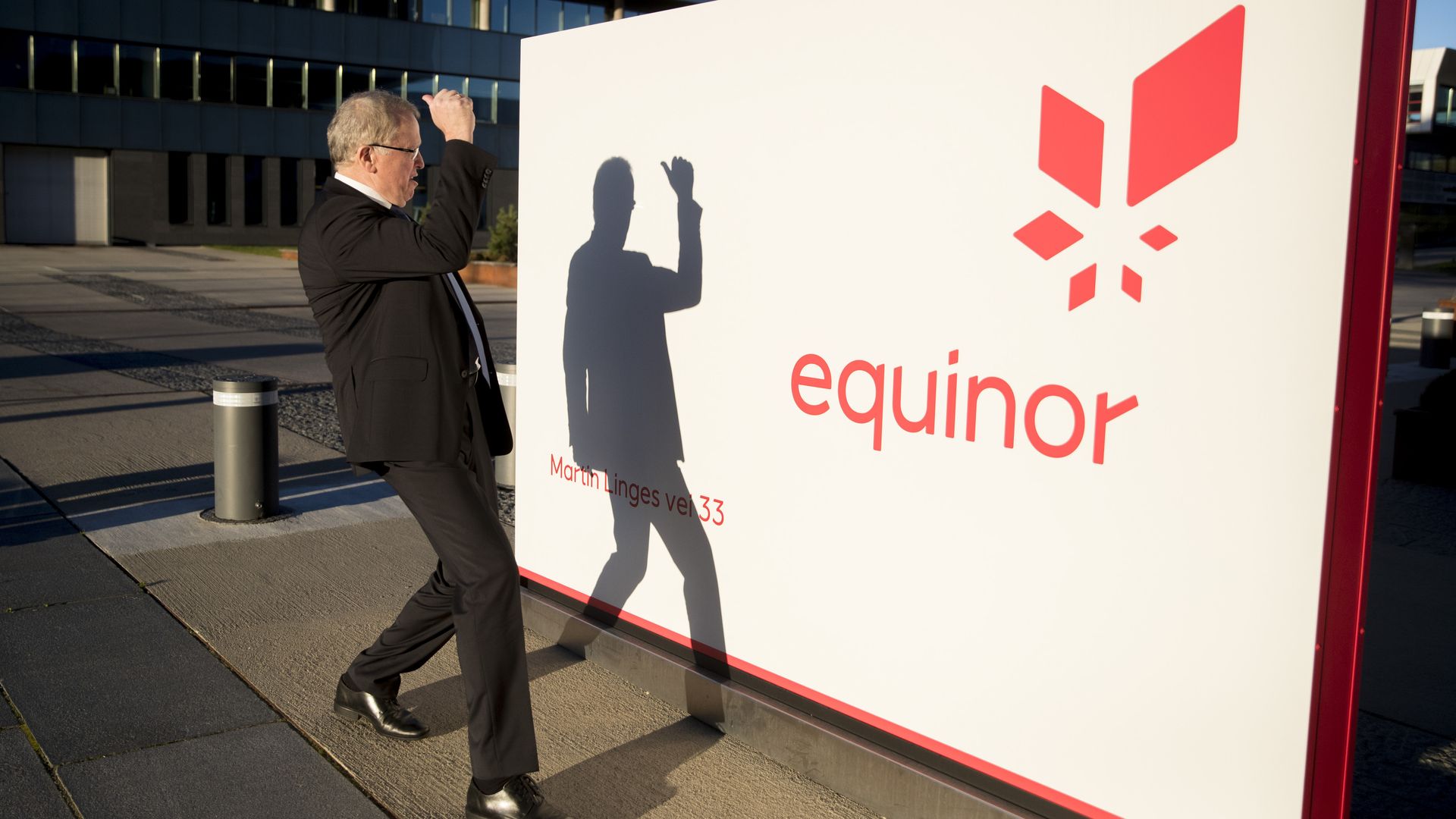 In this image, a suited man shields his face from the sun and looks at his own shadow reflected on a billboard with the Equinor logo on it. 