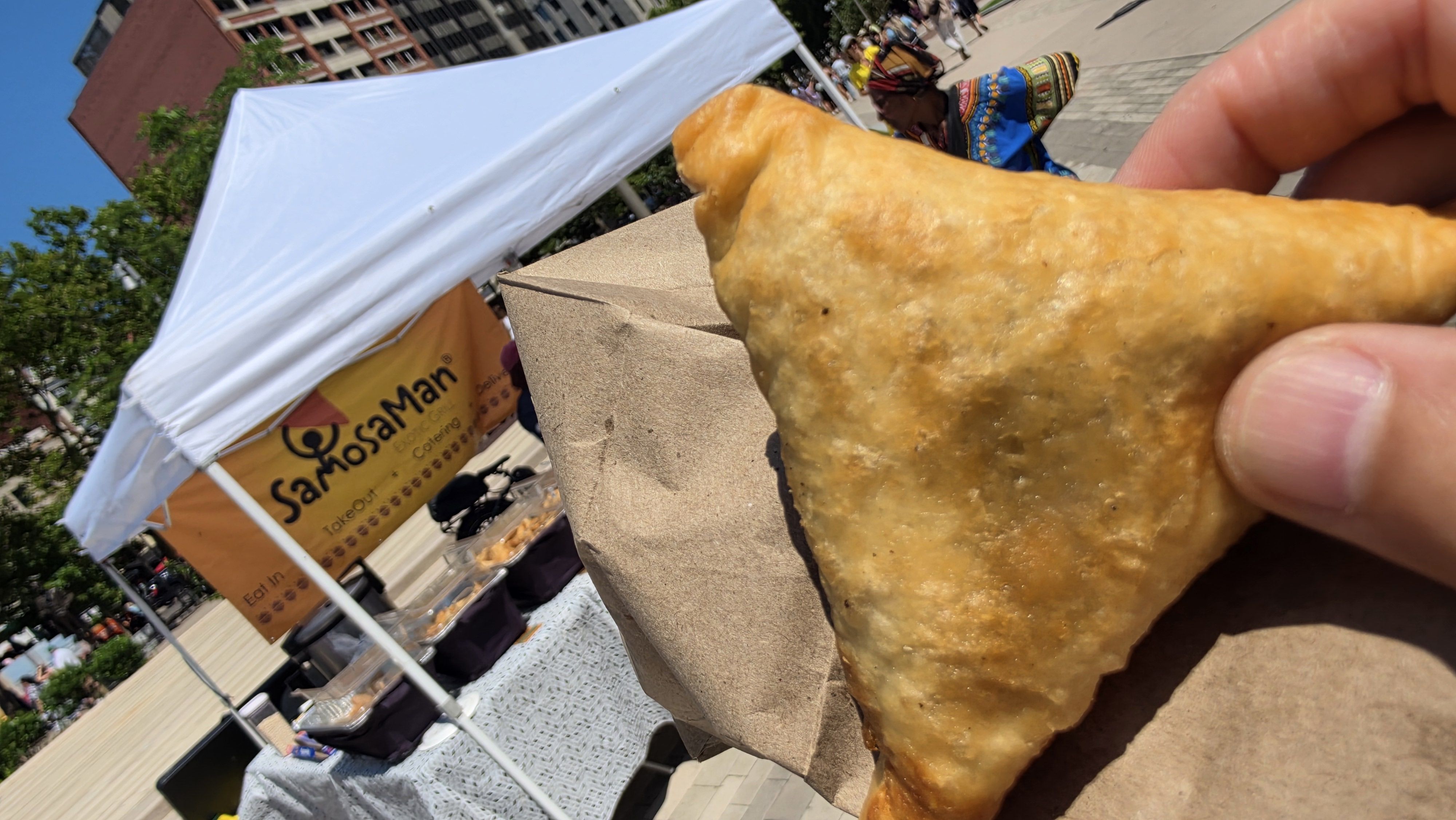 Close-up of a large, golden fried pastry held by a hand at a sunny outdoor festival. A white canopy, a yellow banner reading SamosMan, and people in colorful clothing are visible in the background.