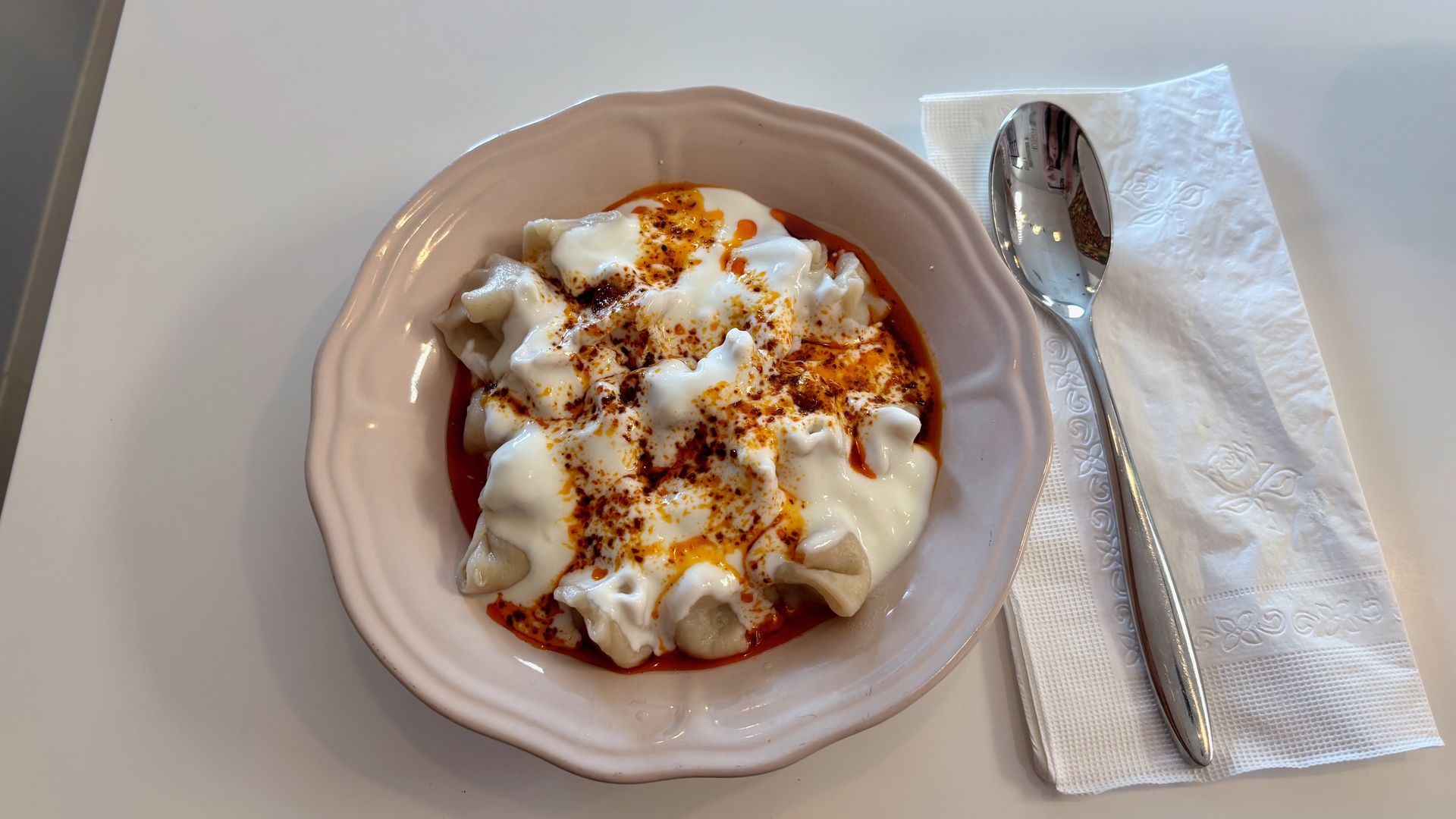 Top-down view of a pale pink bowl of dumplings topped with white yogurt sauce and orange-red chili oil and flakes; a spoon lies on a white napkin to the right.