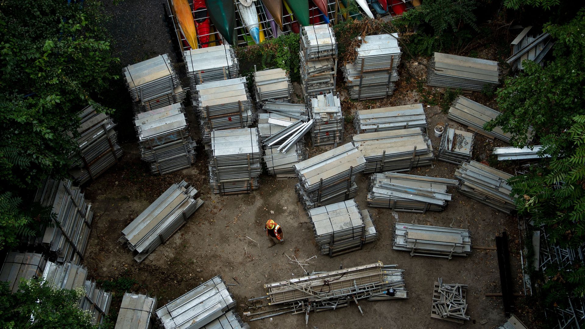 In this image, a construction worker stands in the middle of a clearing surrounded by stacks of steel beams. Trees surround the clearing, and there are also stacks of canoes
