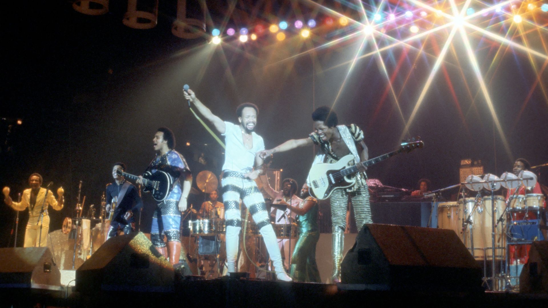 Live concert scene with a band on a dark stage, a lead singer in white raising a microphone while other musicians play guitars and drums beneath bright rainbow spotlights.