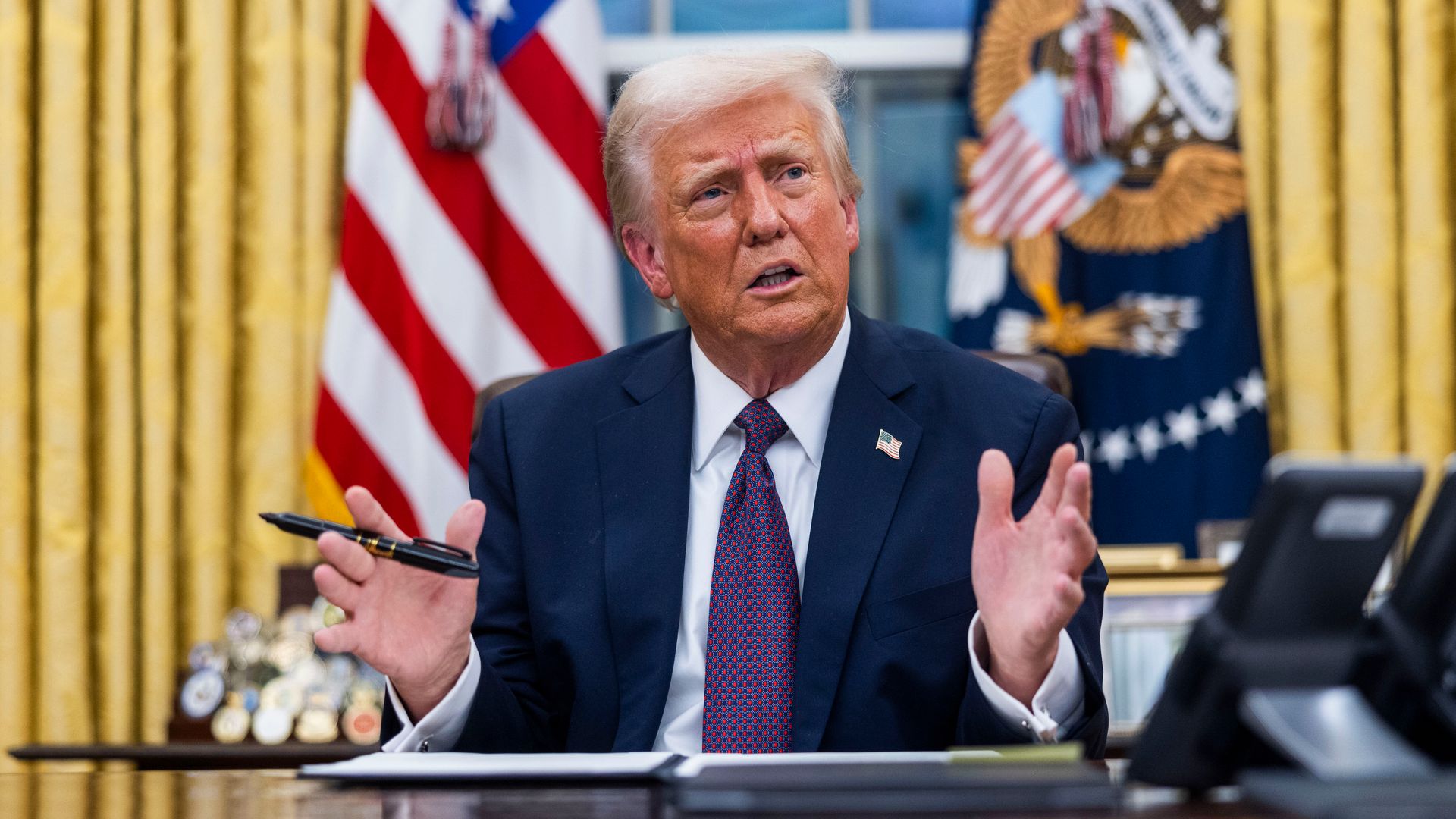 Donald Trump, wearing a blue suit and holding a pen while sitting at the resolute desk in front of gold curtain.
