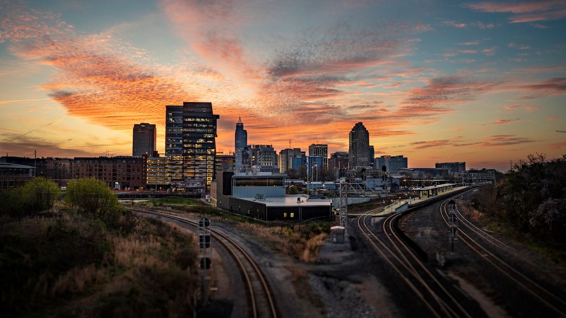 City skyline at sunset with orange and pink clouds, illuminated glass buildings, and two curved train tracks leading into the city. Green trees and shrubs surround the tracks.