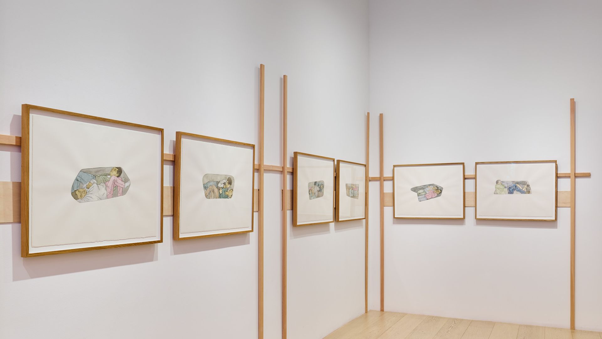 Gallery wall with seven framed watercolor paintings of people resting inside irregular shapes, mounted on white walls with light wooden trim and light wood flooring.