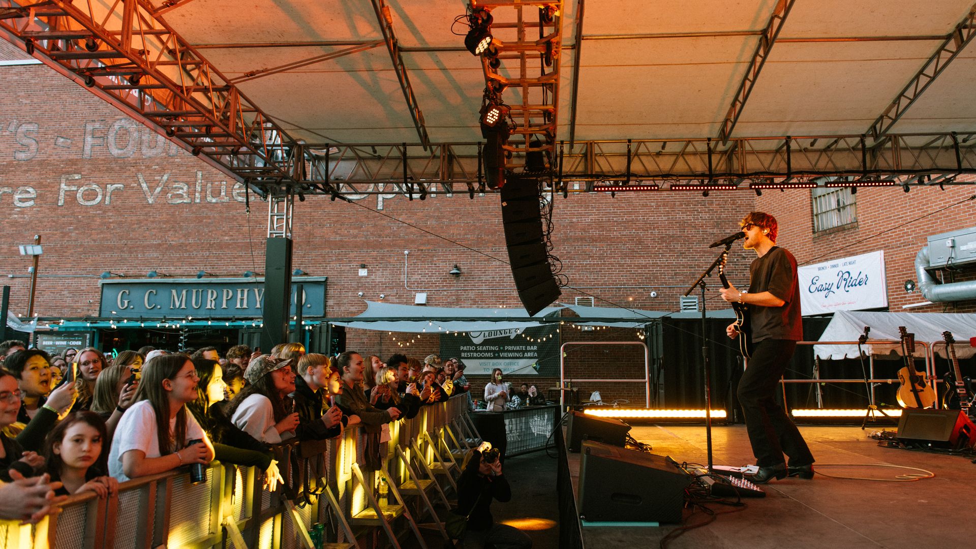 An outdoor concert: a guitarist sings into a mic on a stage with orange lighting and large overhead speakers, as a cheering crowd leans on the barrier; brick buildings and signs in the background.