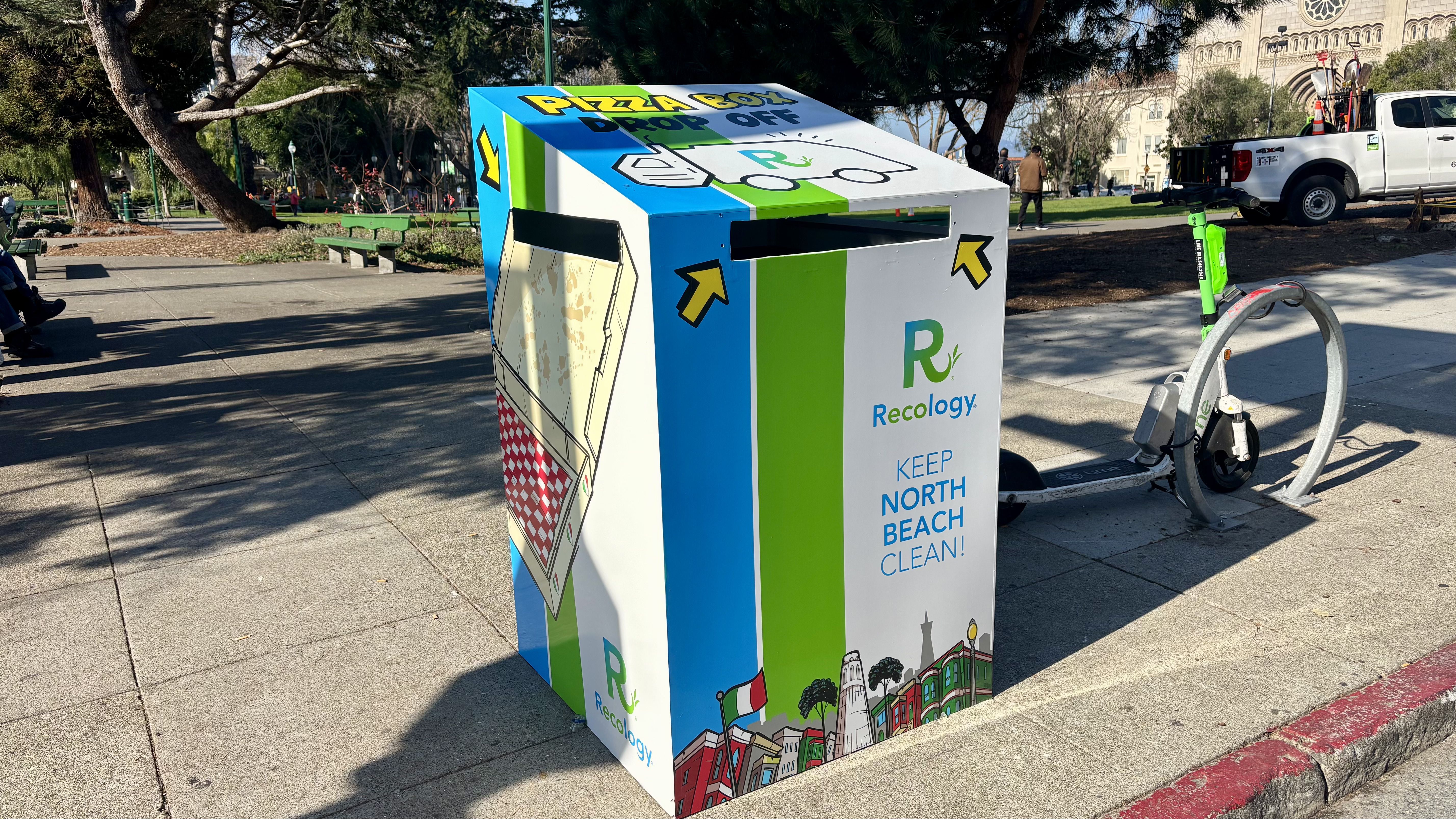 Colorful pizza box drop-off recycling bin with green, blue, and white panels, located on a sidewalk near a green scooter and park benches in a sunny outdoor setting.