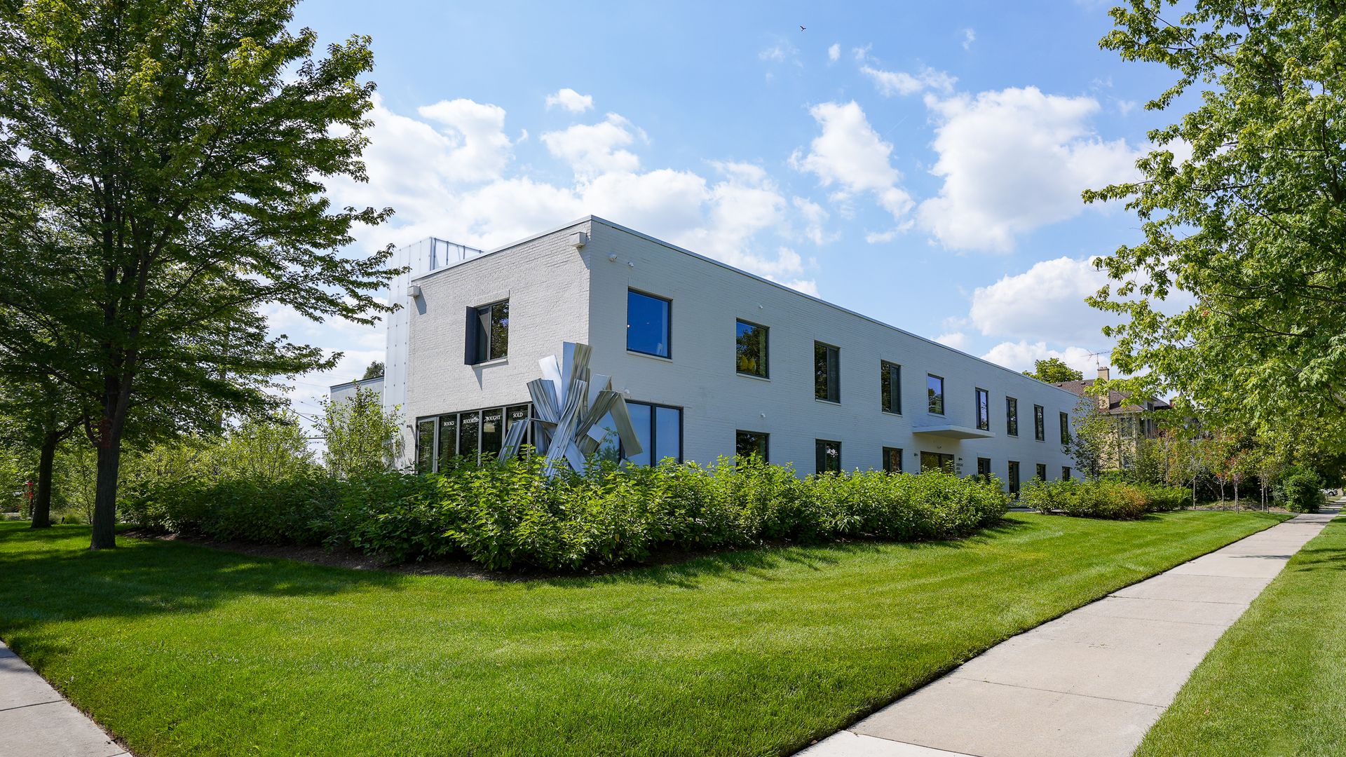 Sunlit white modern building with large windows, surrounded by green lawn and trees. A metal sculpture and curved sidewalk frame the foreground, under a blue sky with white clouds.