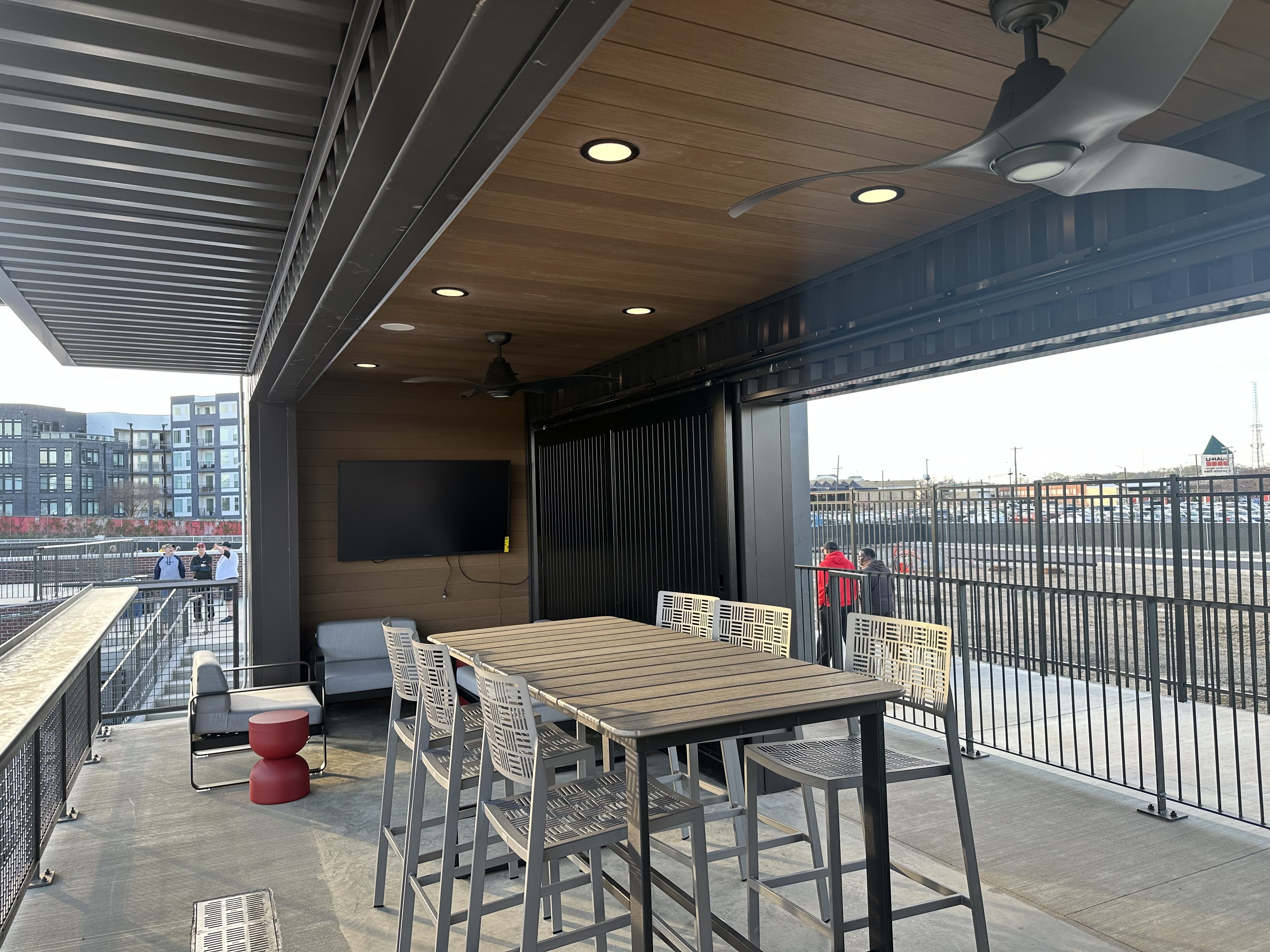Outdoor covered patio with a long wooden table and gray woven chairs, a wall-mounted flat-screen TV, ceiling fans, and recessed lights. A railing overlooks a rail yard and distant buildings.