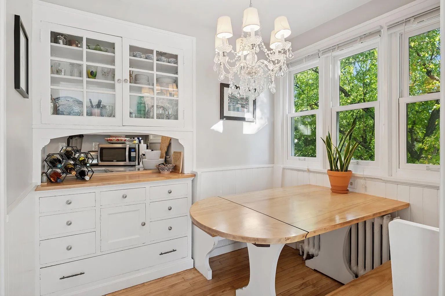 Bright white kitchen corner with wooden table, crystal chandelier, built-in cabinet with dishes, wine rack, microwave, and large windows showing green trees outside.