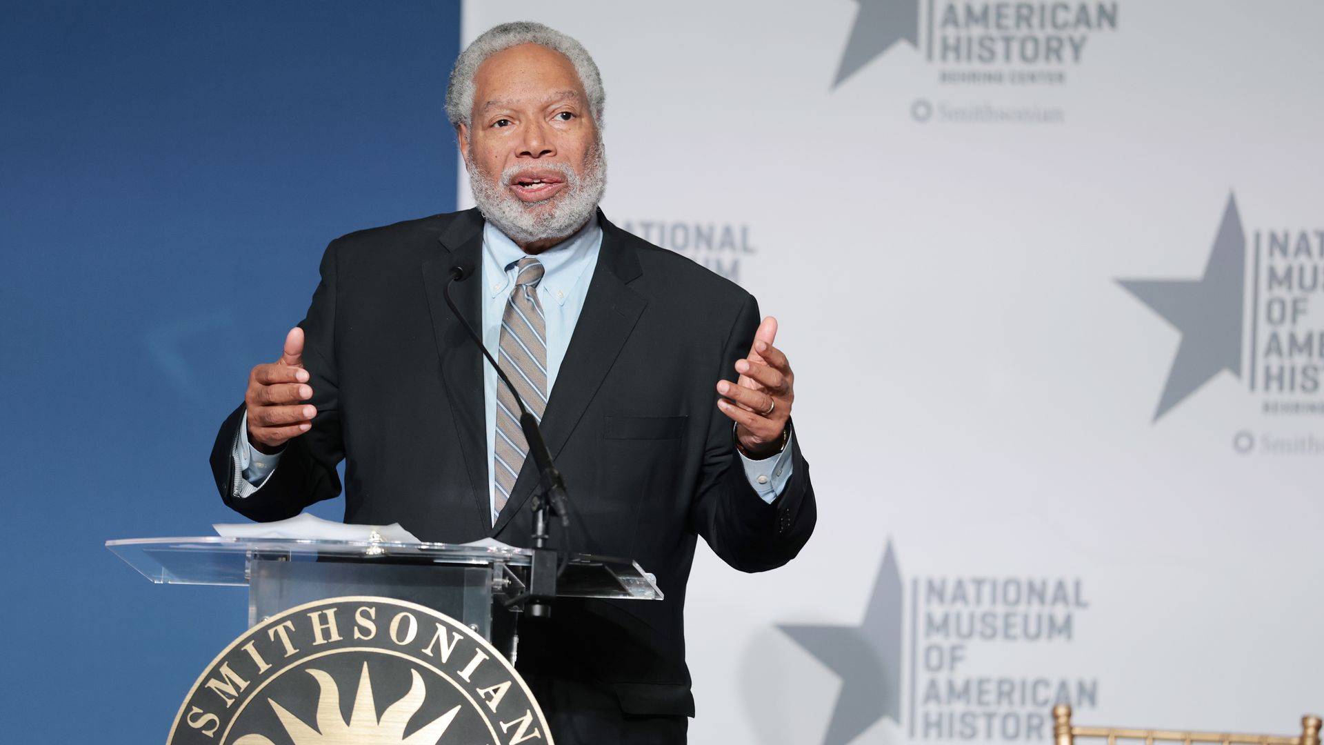 An older man with a gray beard wearing a dark suit and striped tie speaks at a podium with a Smithsonian seal; blue backdrop and a large "National Museum of American History" sign in the background.