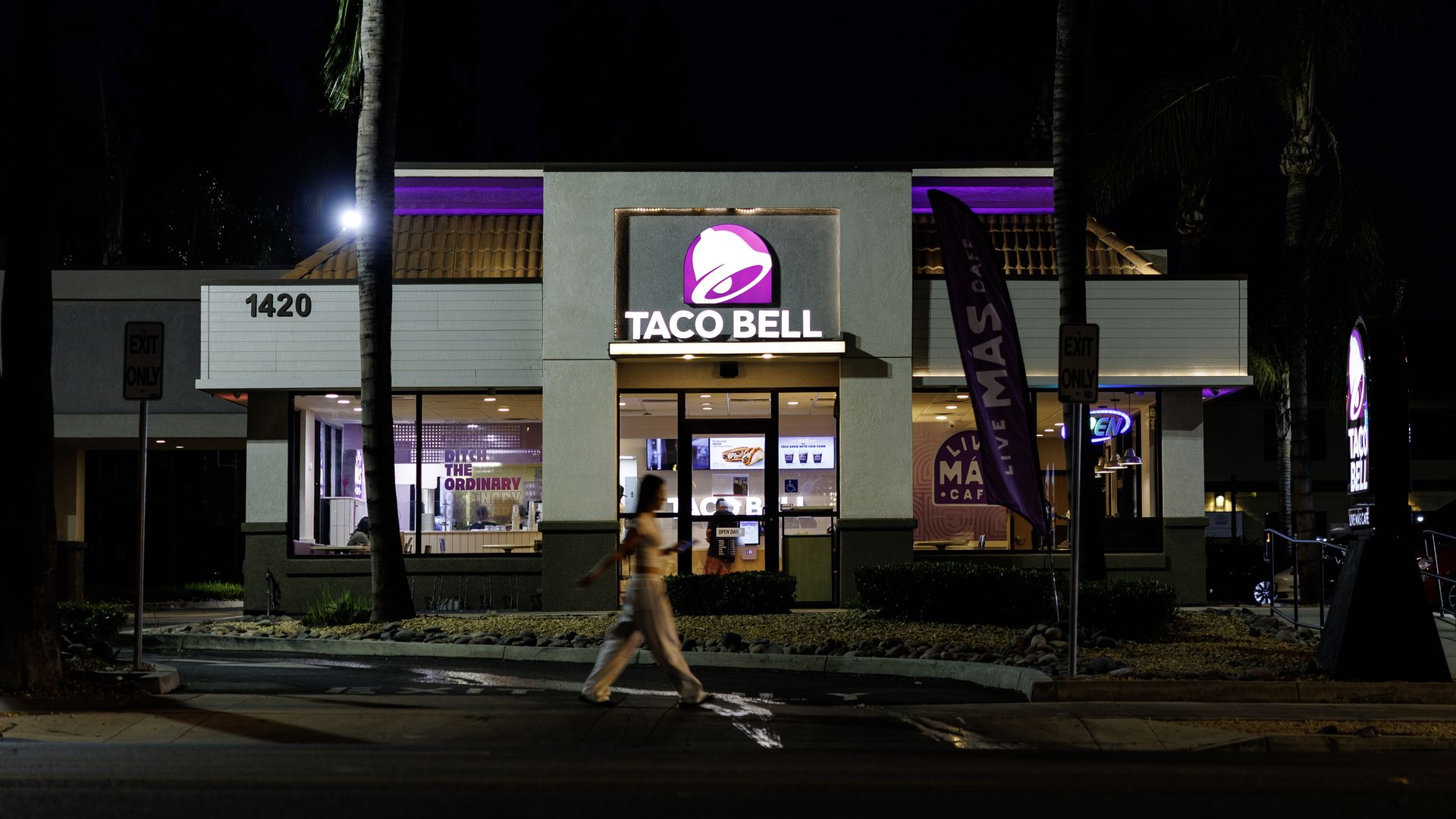 Night view of a Taco Bell restaurant with purple neon lighting, palm trees flanking the gray facade, a passing blurred figure in the foreground, and a bright Taco Bell sign centered above the entrance.