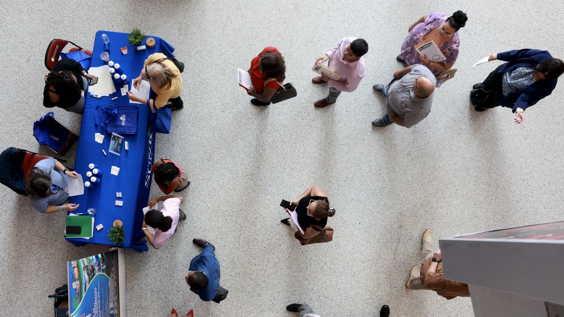 Lines of people, seen from overhead, in front of a blue table covered in paperwork. 