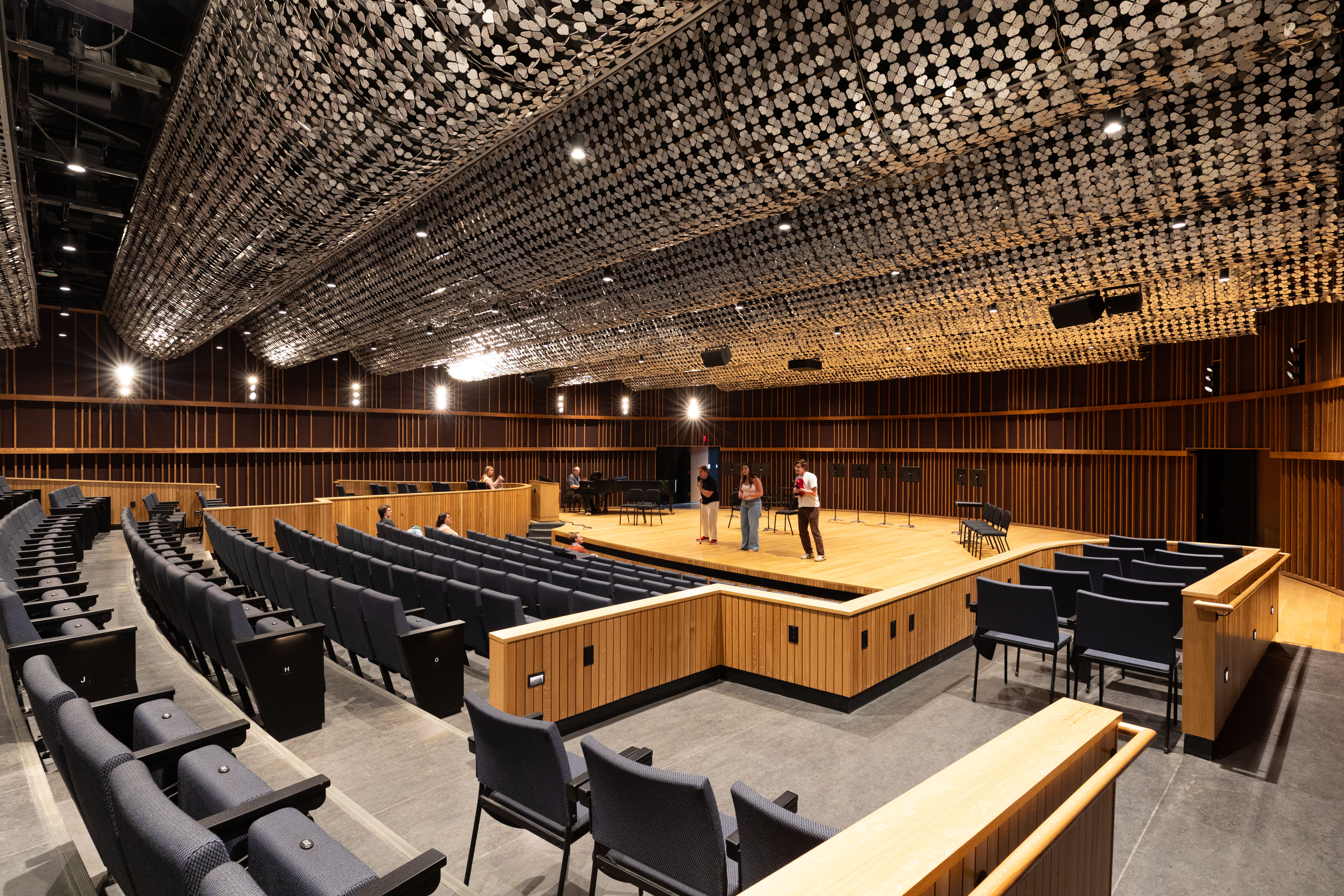 A modern concert hall with tiered seating and a wooden stage, where a small group rehearses under a textured ceiling.