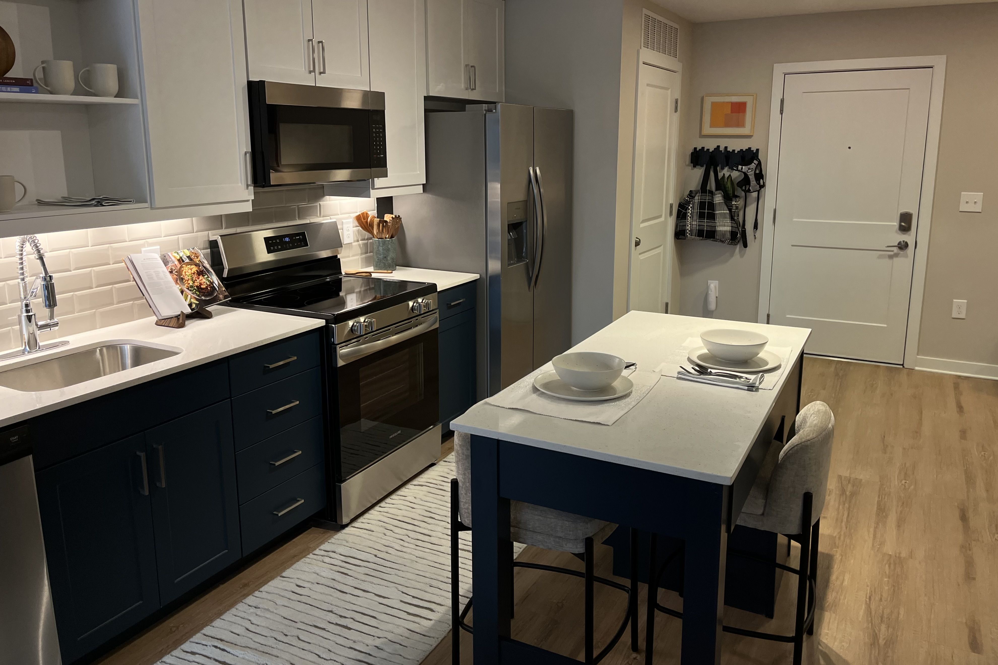 Modern kitchen with navy lower cabinets, white upper cabinets, stainless steel appliances, white countertop island with two place settings, light wood floor, and beige walls with a white door.