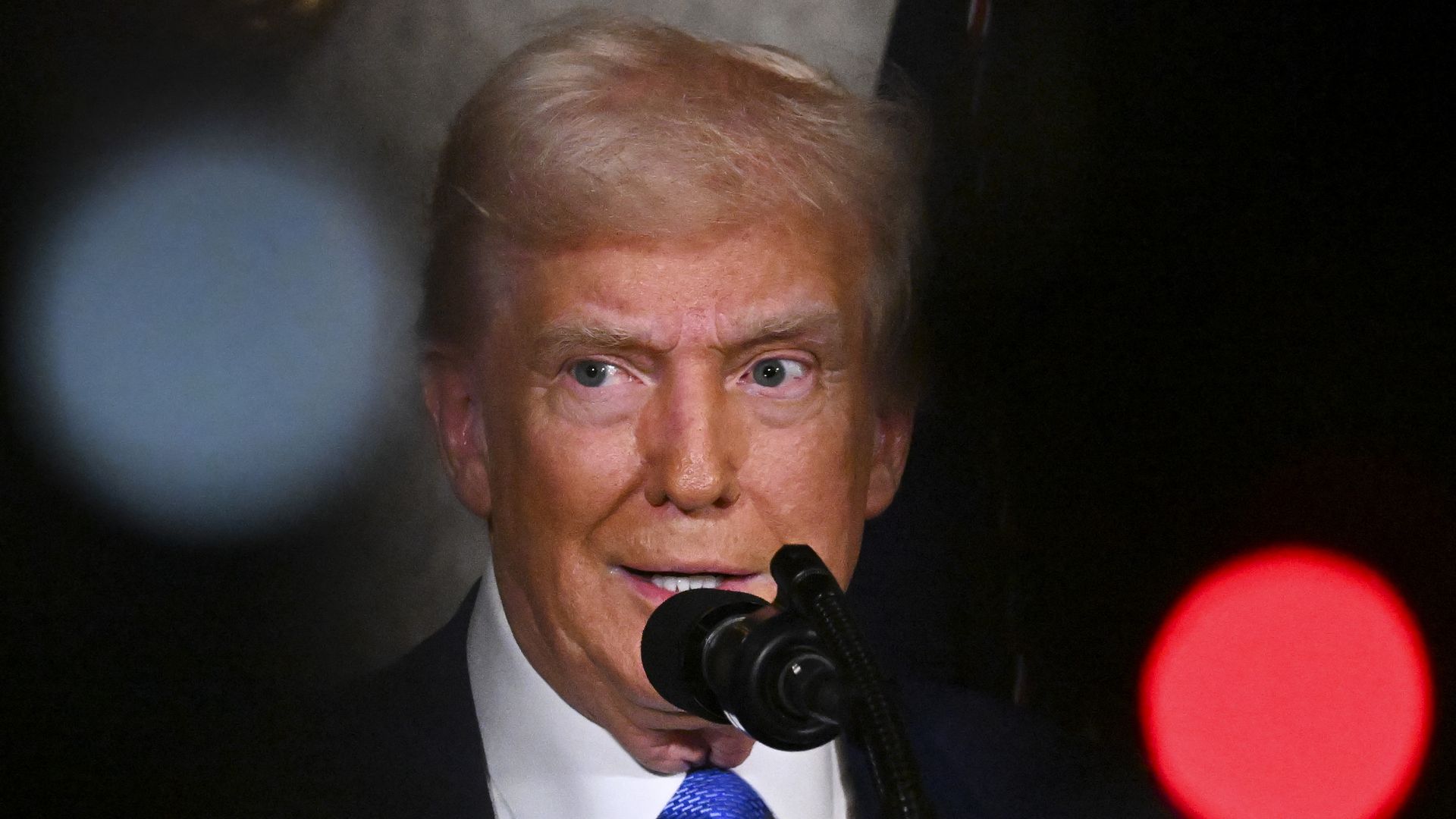 Donald Trump speaks at a microphone wearing a black suit while surrounded on both sides by dark shadows.