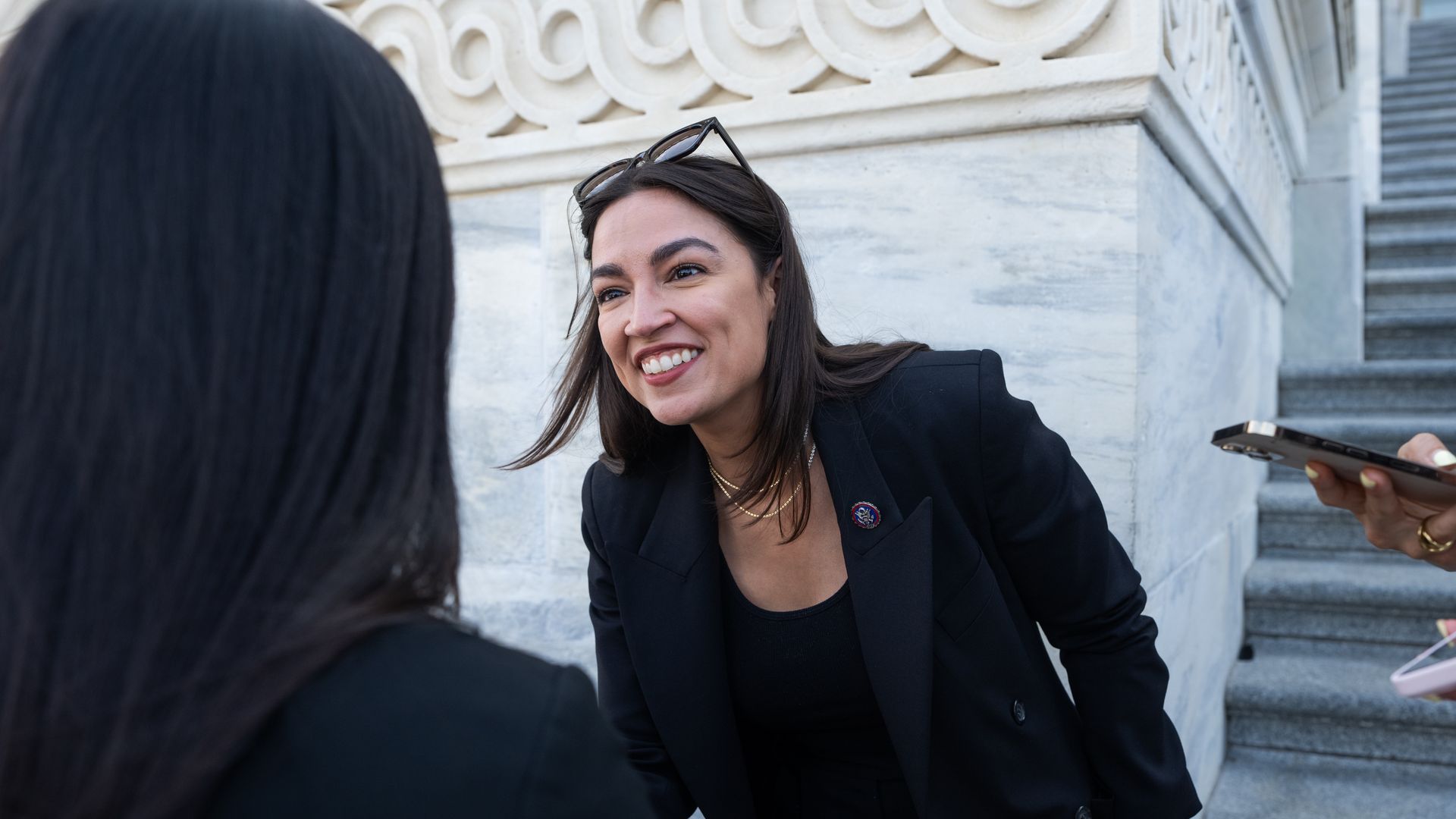 Alexandria Ocasio-Cortez leans forward to talk with someone in the foreground outside the Capitol.