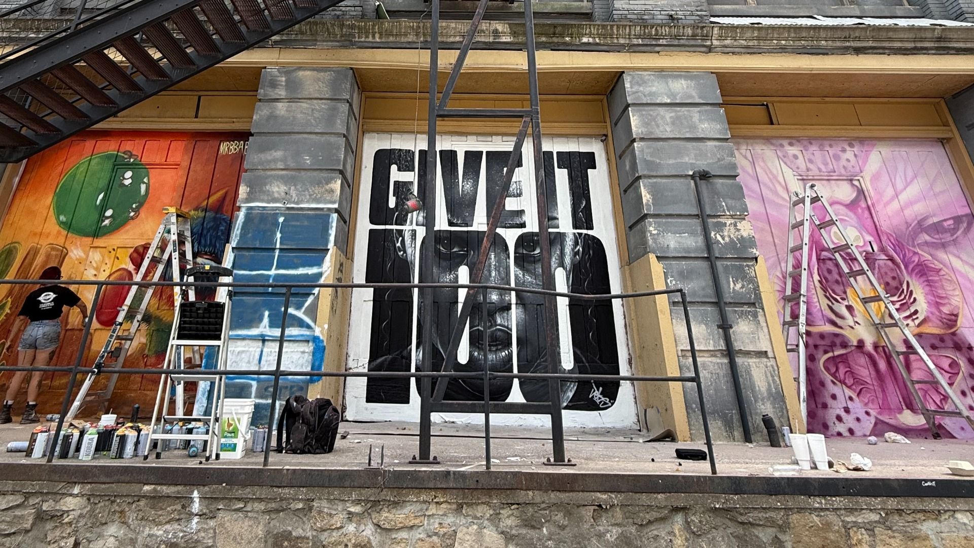 Urban storefront mural with bold "GIVE IT UP" on a white center panel, flanked by orange left and purple right murals. Ladders, spray cans, a painter on the left, and metal stairs overhead.
