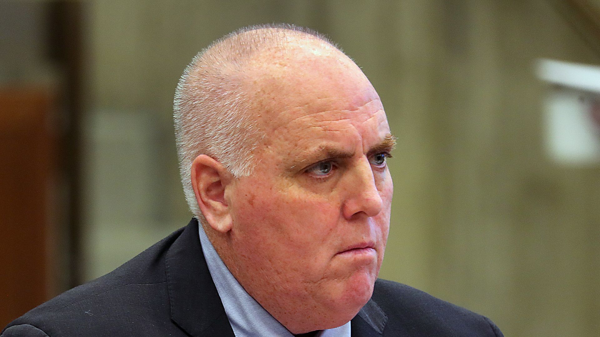 Serious bald man in a black suit, light blue shirt, and red tie with white dots, looking to the side against a blurred indoor background.