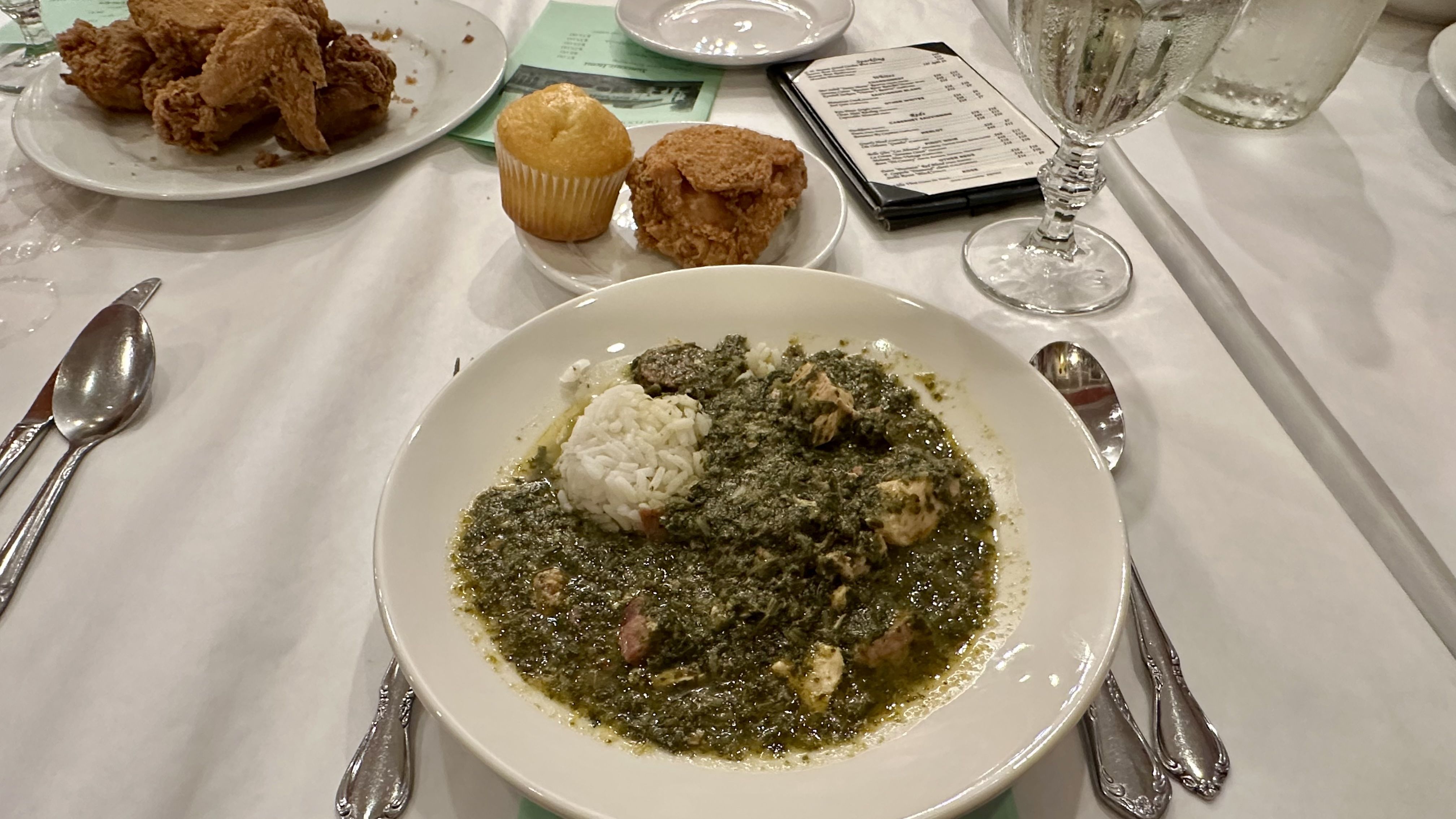 A bowl of gumbo on a table set with fried chicken, a muffin, menu and a water glass in the background.