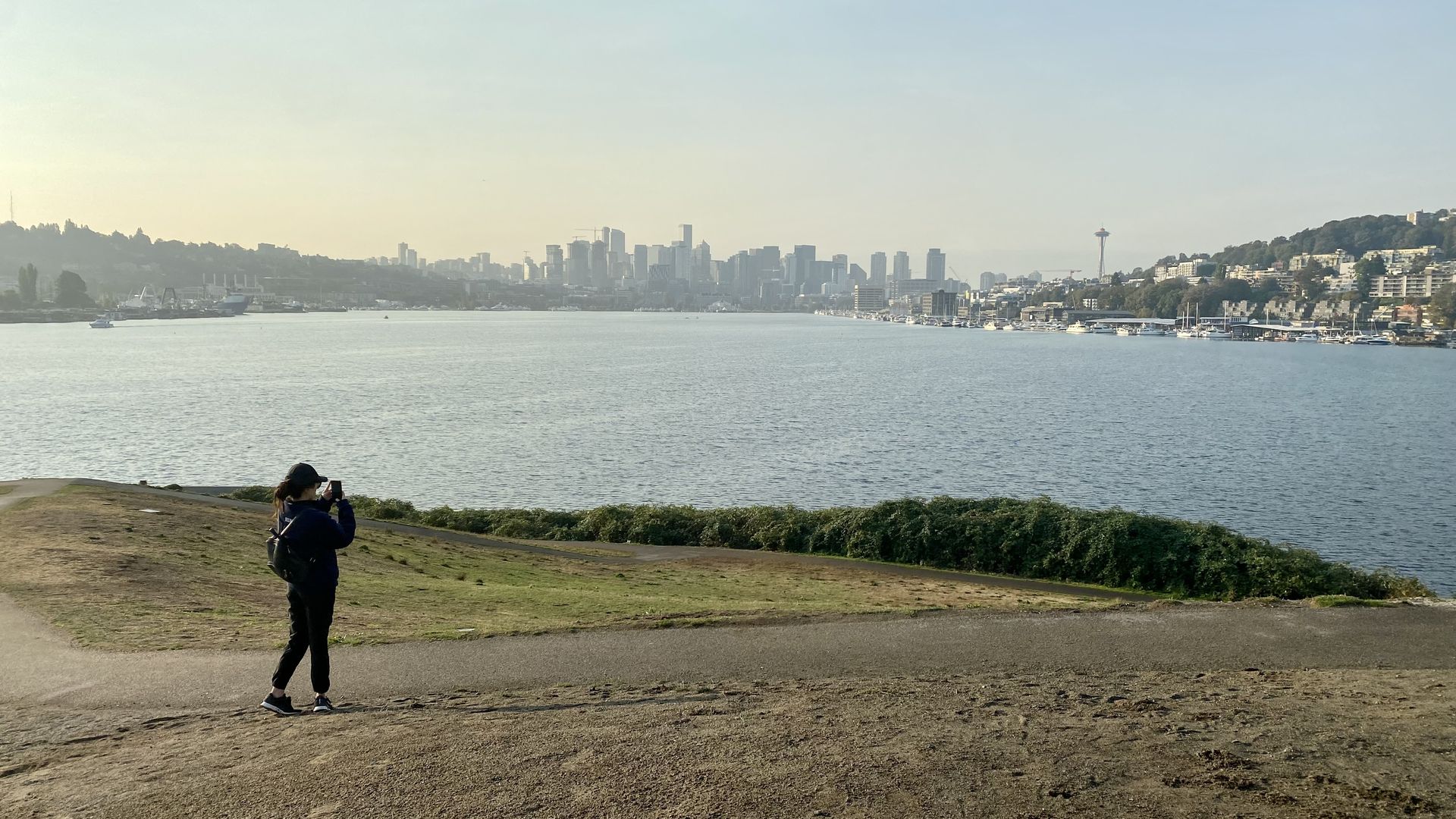 A person takes a photo of the smoky Seattle skyline from a hill on a lake.