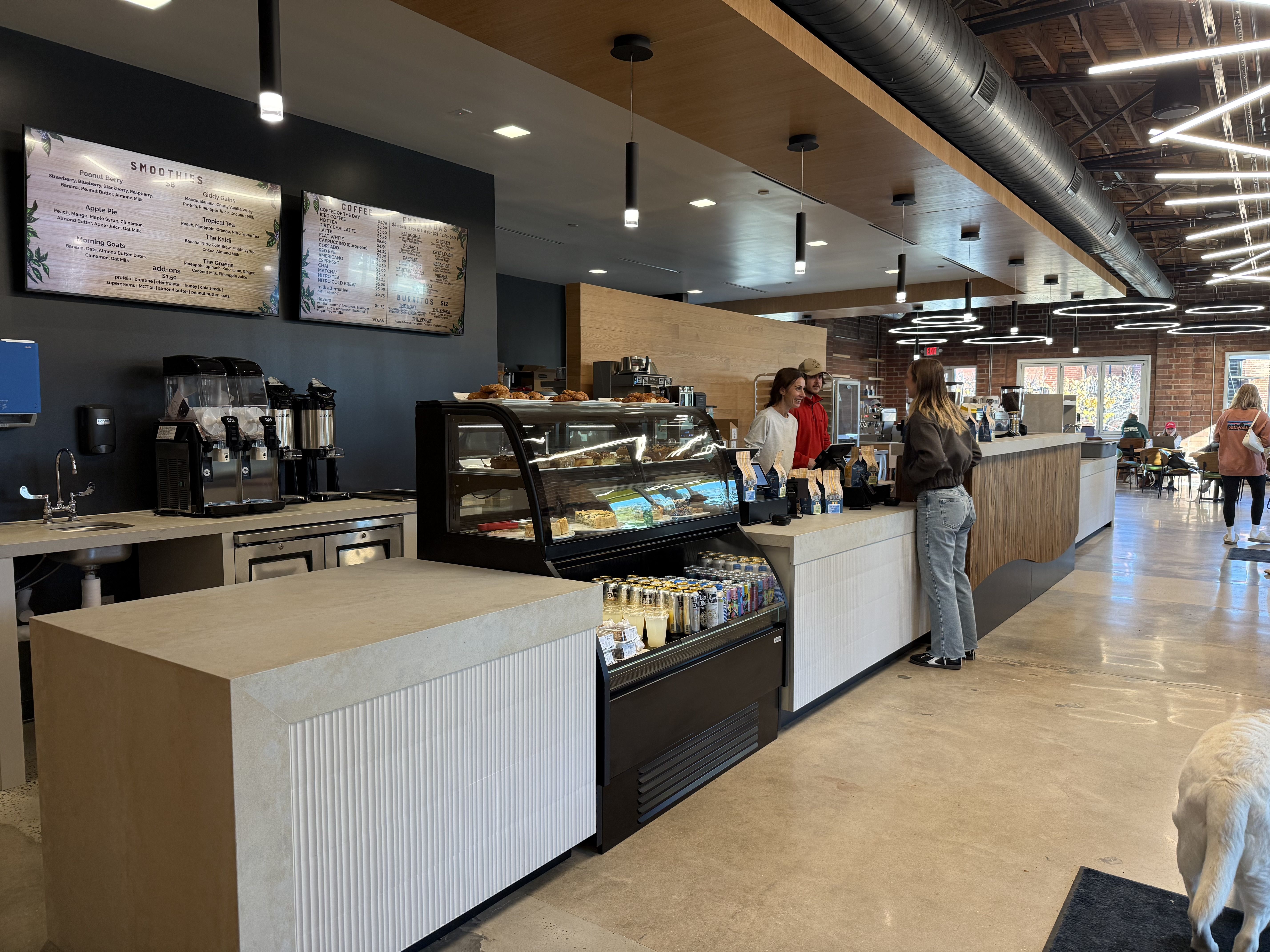 Modern cafe interior with light wood and black accents, a display case of pastries and drinks, digital menu boards, and customers ordering at the counter under circular ceiling lights.