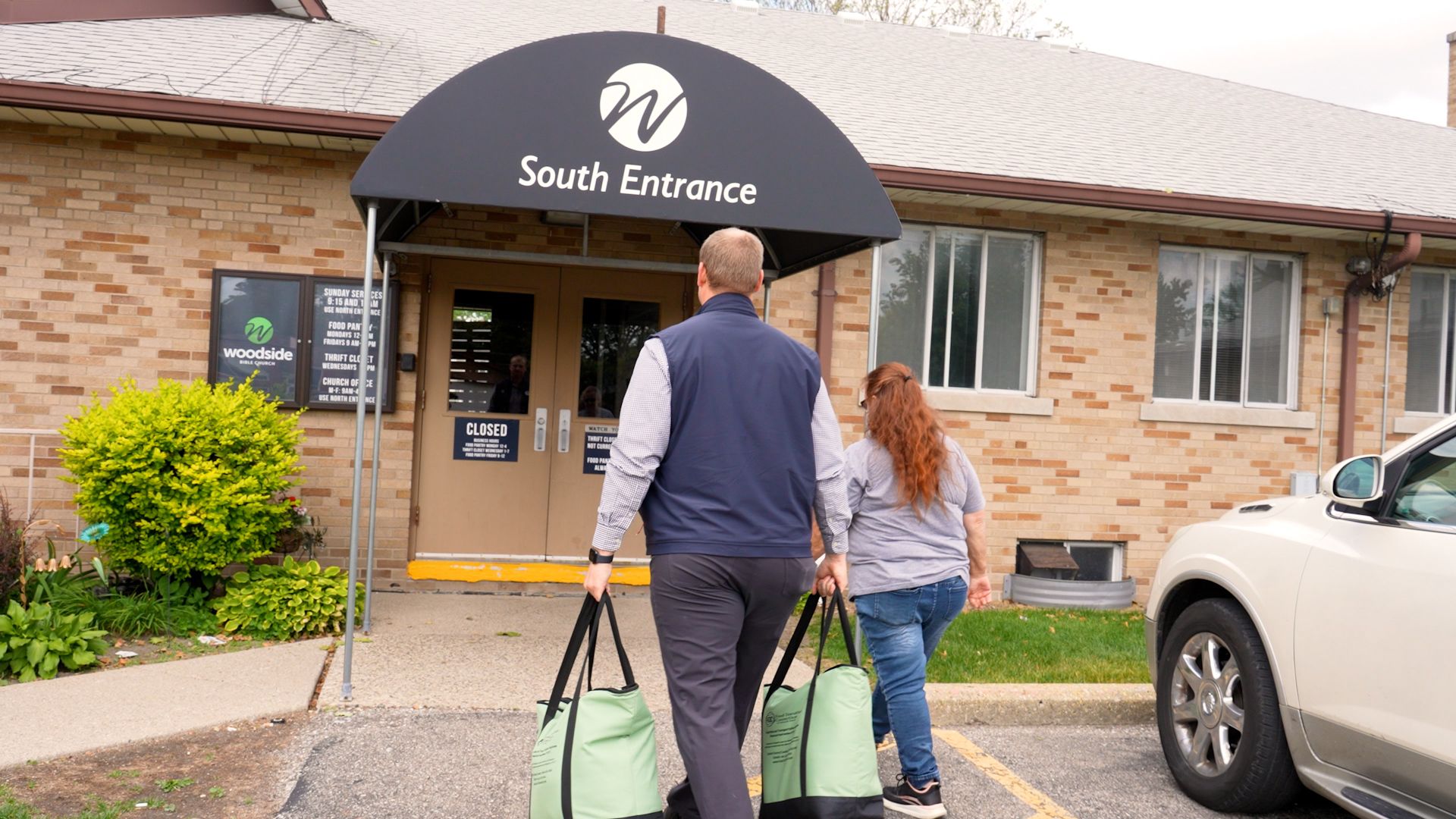 Two people carrying green bags enter the South Entrance of a brick building labeled Woodside Bible Church, with a white vehicle parked nearby and plants by the sidewalk.