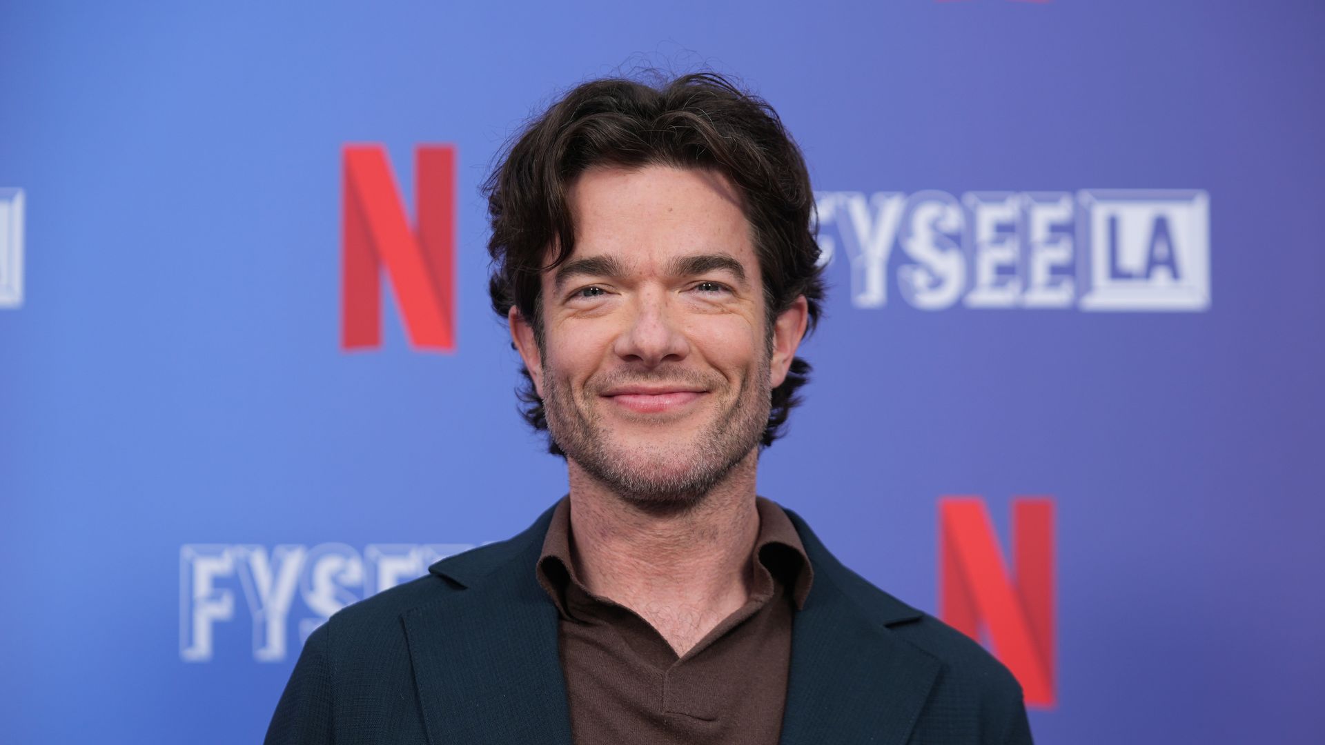 Smiling man with dark wavy hair and light beard wearing a dark blazer over a brown shirt stands against a blue backdrop with red Netflix logos and white FYSEE LA text.
