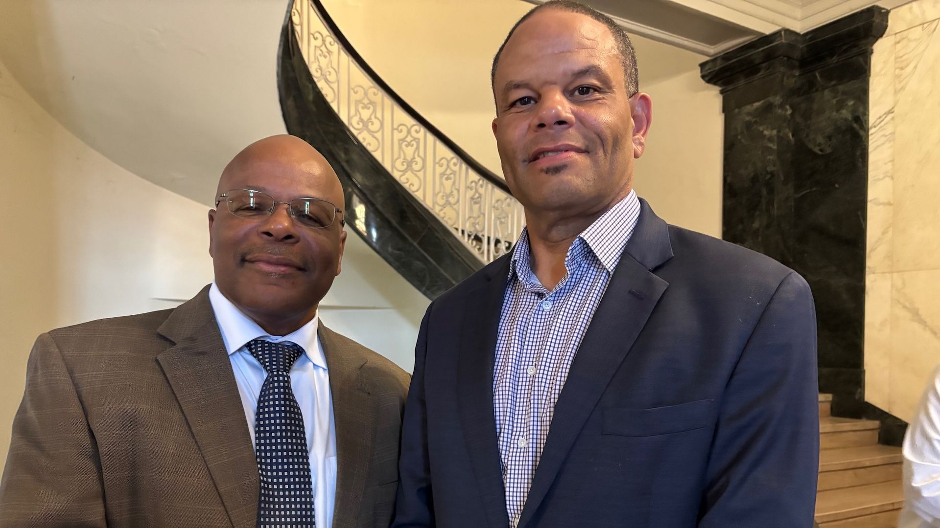 Two men in business suits pose in a marble lobby beside a curved staircase with ornate railing. Left man wears brown suit and glasses; right man wears navy suit with a checkered shirt.
