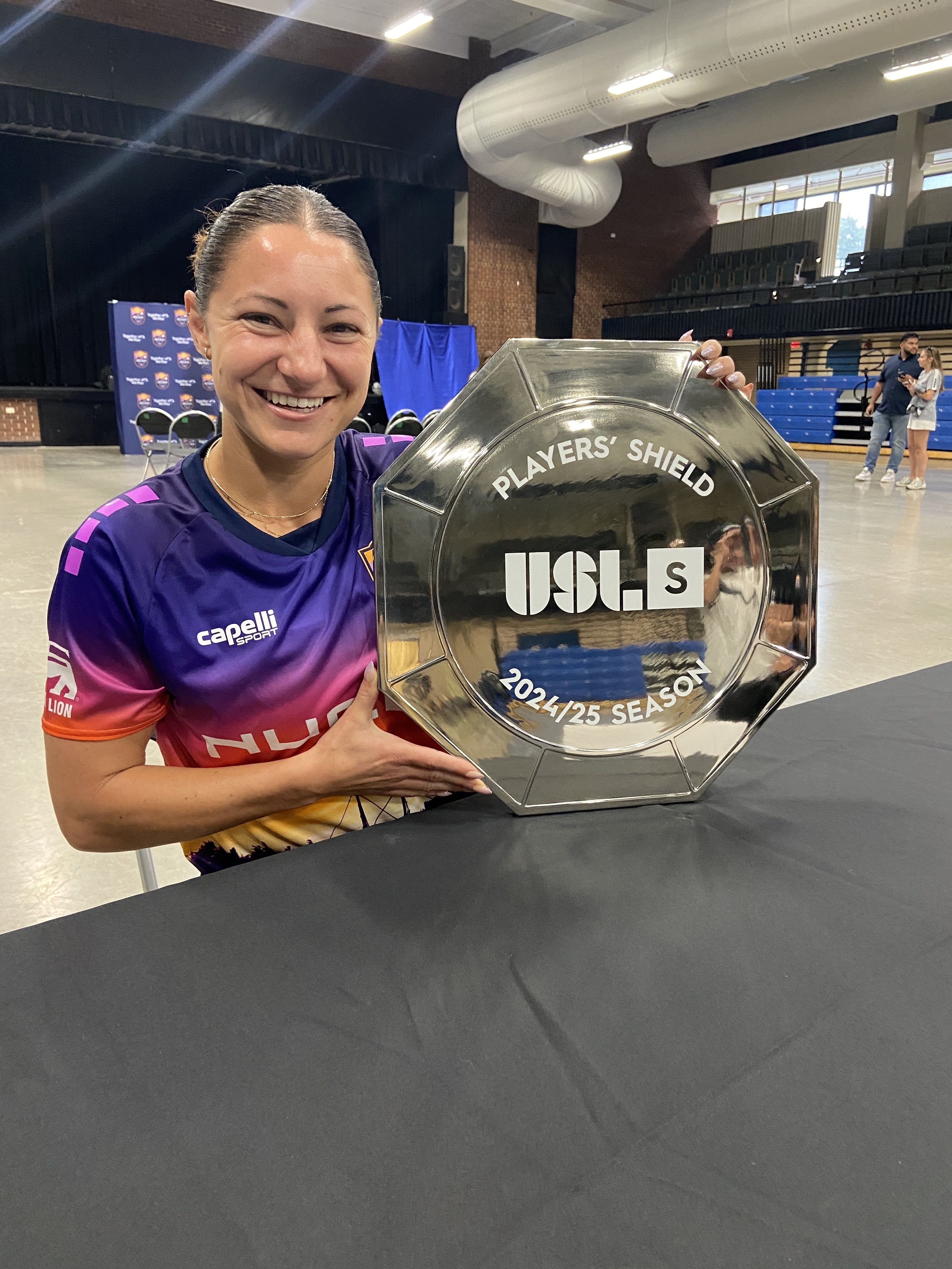 Woman in purple and pink sports jersey smiling and holding a shiny octagonal Players' Shield trophy for the 2024/25 USL season indoors on a table.