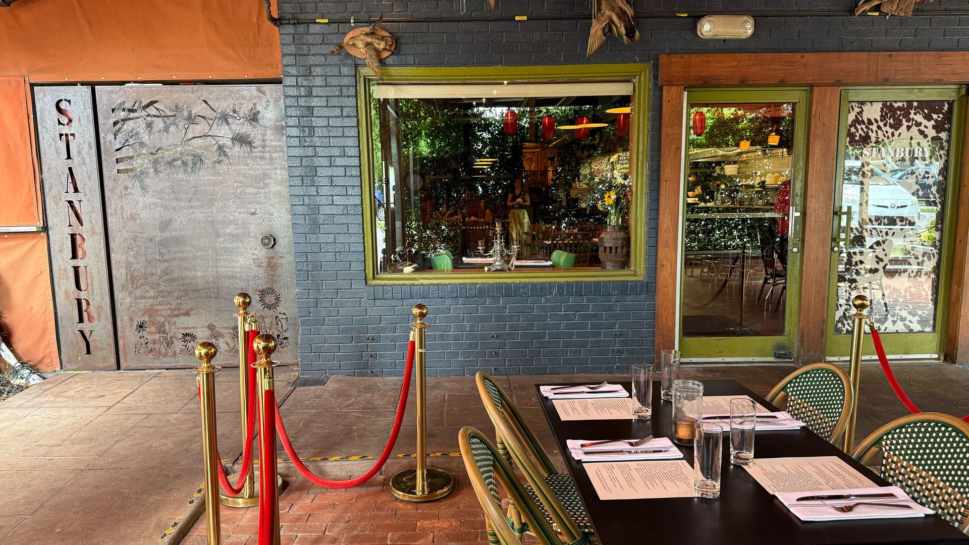 Outdoor dining area of Stanbury restaurant with a table set for four, green woven chairs, red lanterns hanging, black brick wall, and a metal door with Stanbury text.