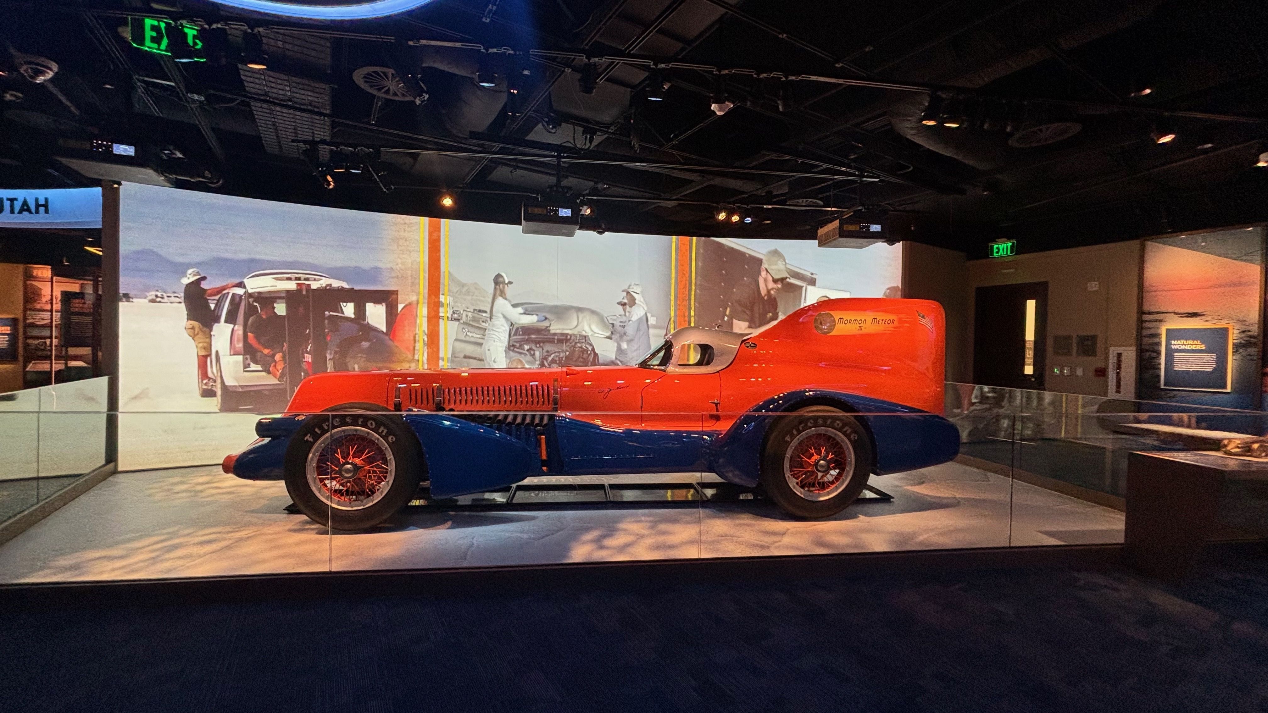 Orange and blue vintage race car labeled "Mormon Meteor III" on display in a museum, with a desert-themed mural, glass barrier, and a dark ceiling with overhead lights.