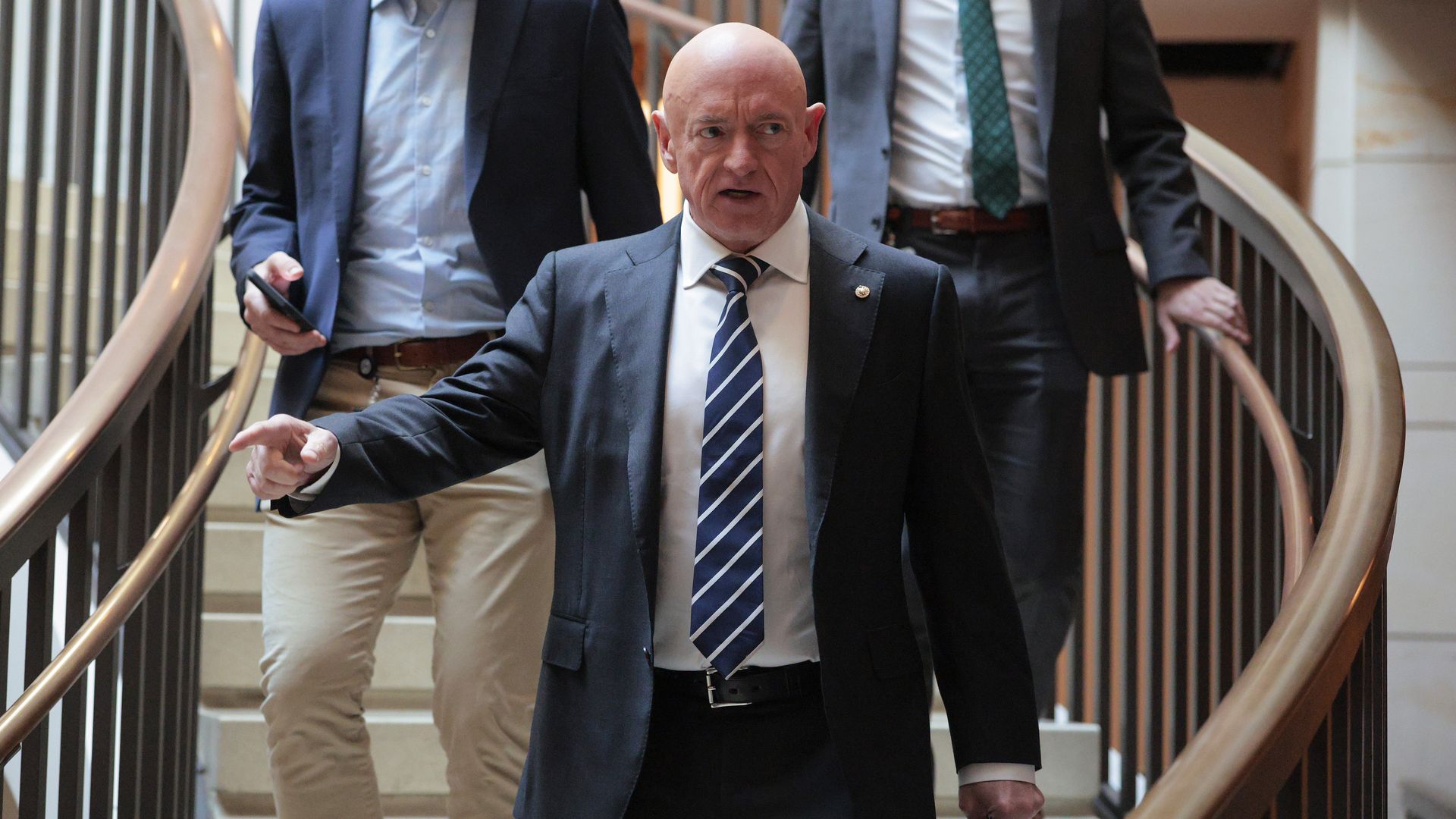 Bald man in a dark suit with a striped tie gestures while walking down a curved staircase, followed by two men in suits, one holding a phone.