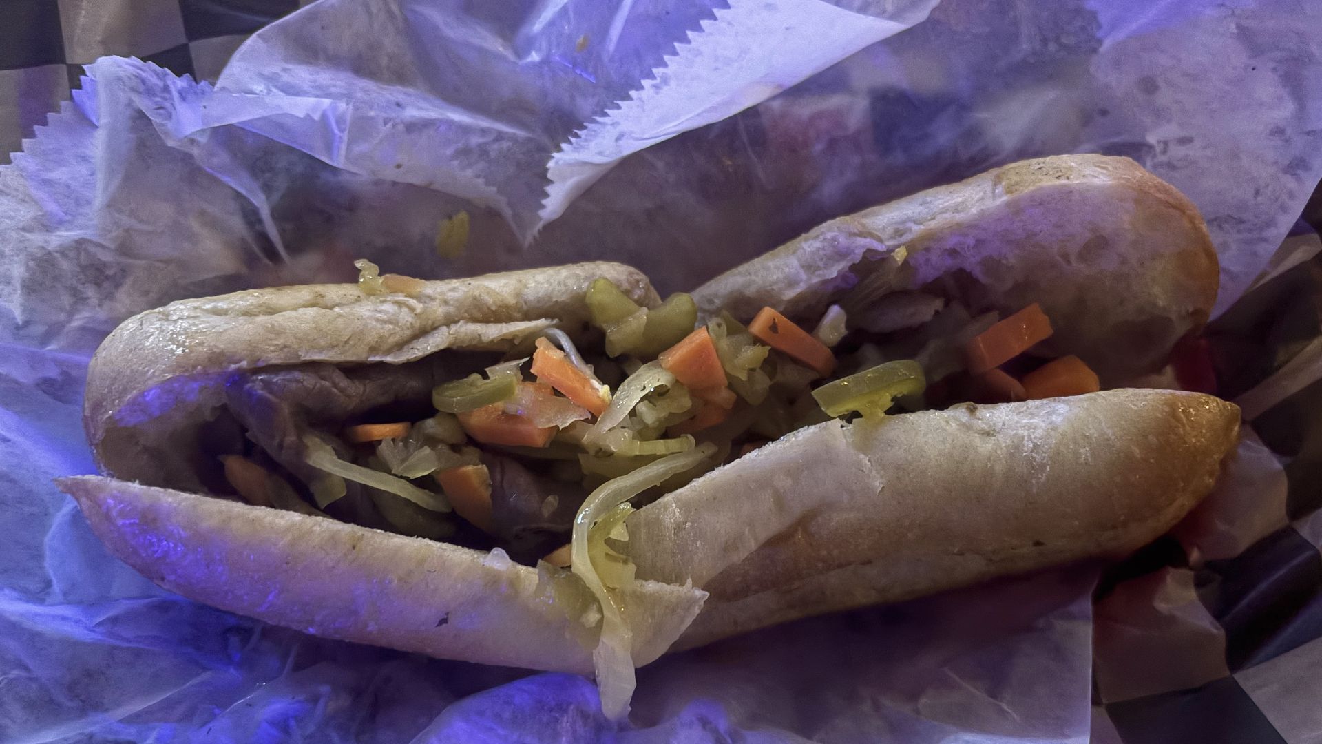 Close-up of a sandwich or wrap filled with diced carrots, celery, and other vegetables in sauce, tucked in crinkled white paper; warm buns and purple-tinted lighting.